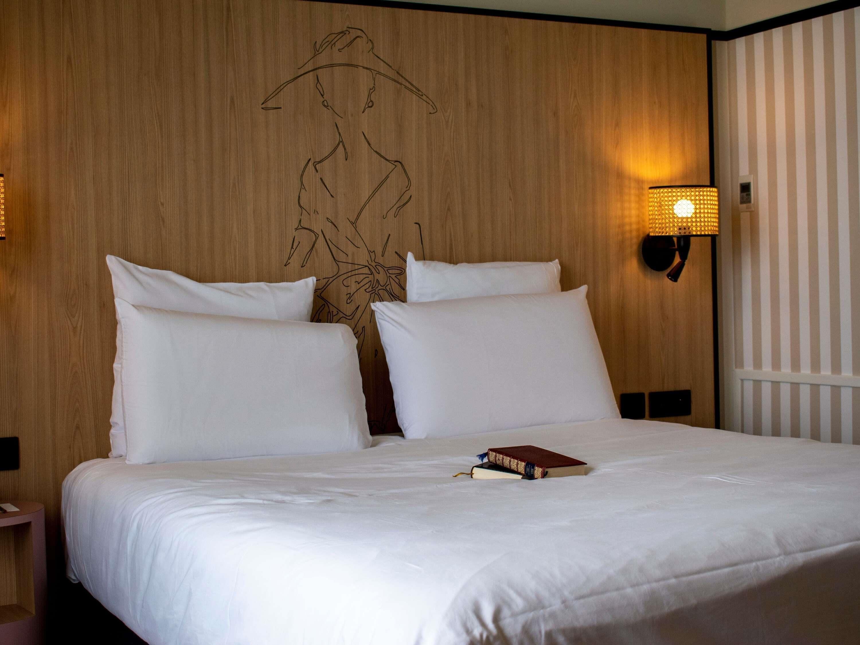 A hotel room with a wooden headboard, white bedding, a bedside lamp, and a book on the bed.