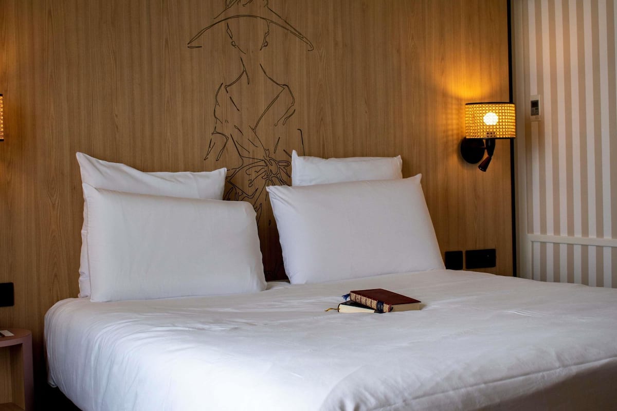 Habitación de hotel con cabecera de madera, ropa de cama blanca, lámpara de noche y un libro sobre la cama.
