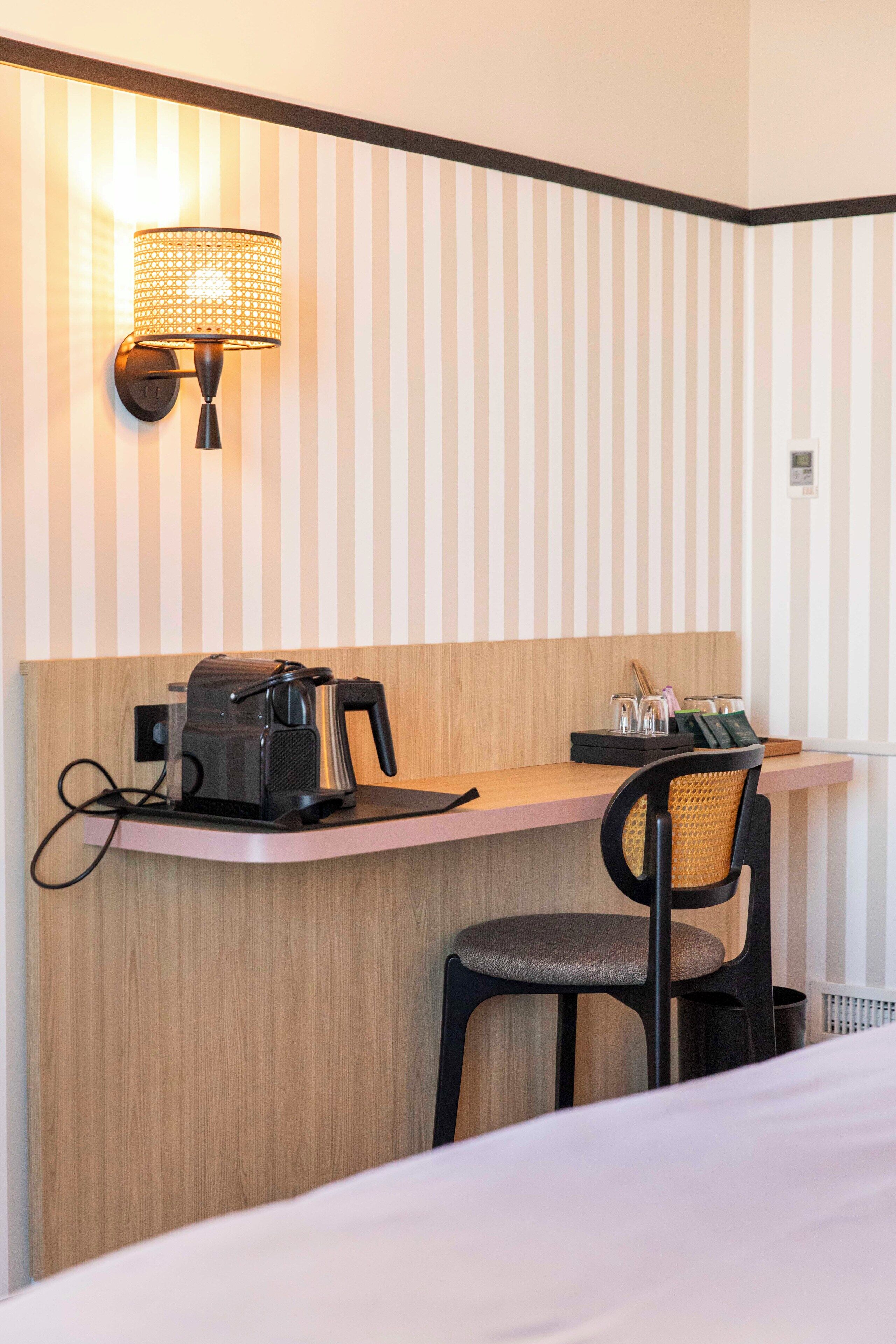 A hotel room with a wooden desk, a chair, a coffee maker, and a wall-mounted lamp.