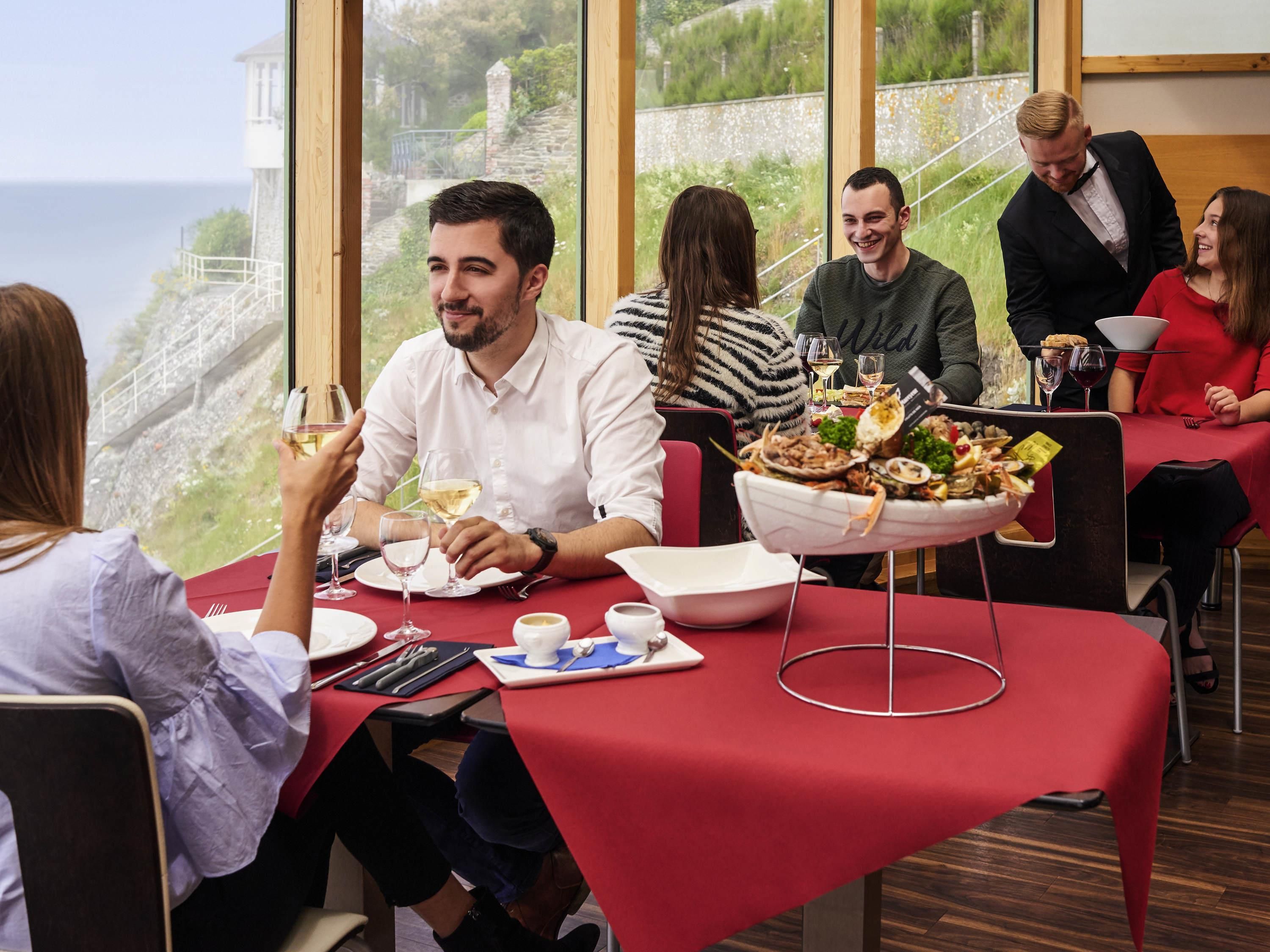 A dining area with tables set for a meal, a view of the sea, and a stone wall in the background.