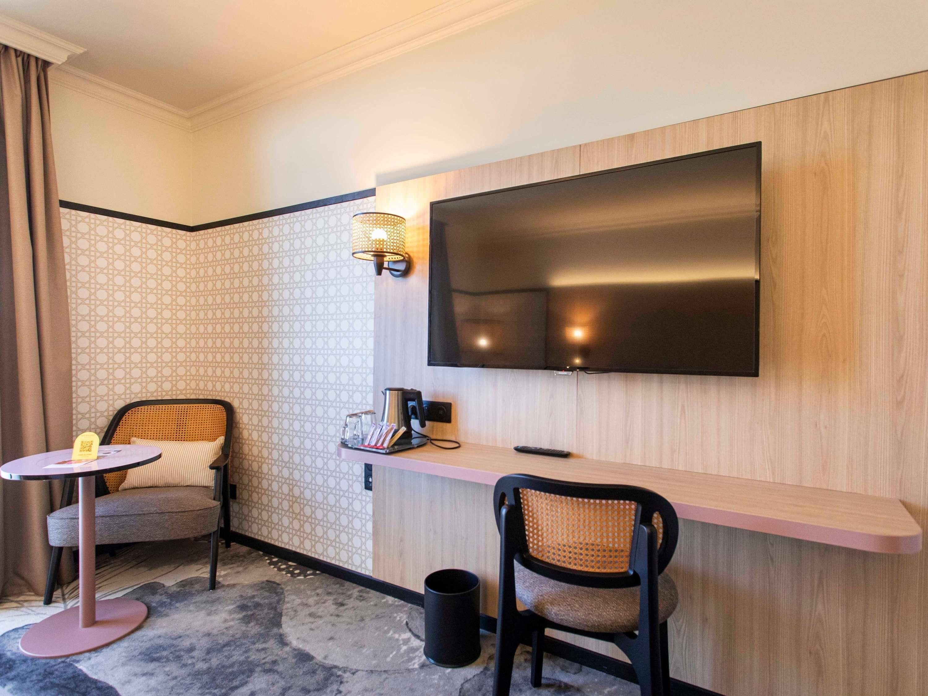 Habitación de hotel con televisión montada en la pared, una pequeña mesa redonda con una silla, otra silla y una mesita con una tetera y un control remoto.