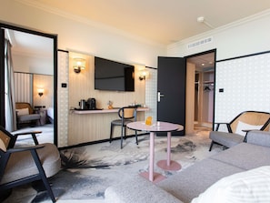 A hotel room with a bed, a seating area with chairs, a small table, a TV mounted on the wall, and a kitchenette with a coffee maker.
