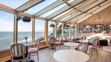 A restaurant with a view of the ocean, wooden interior, and round tables.