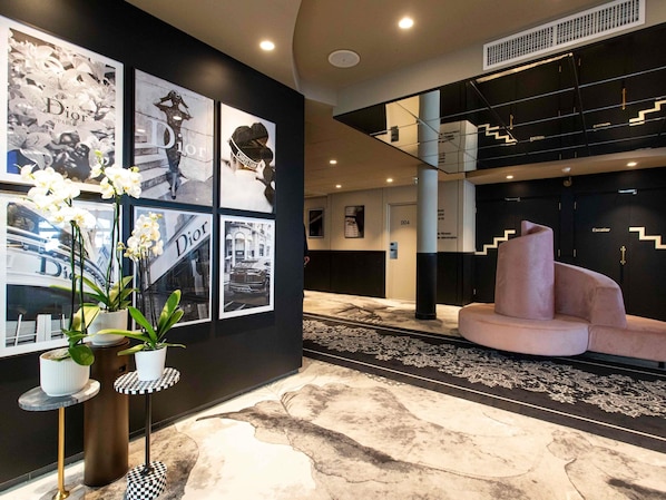 A lobby with a black wall adorned with framed pictures, a large potted orchid plant, a checkered table, and a pink sofa.