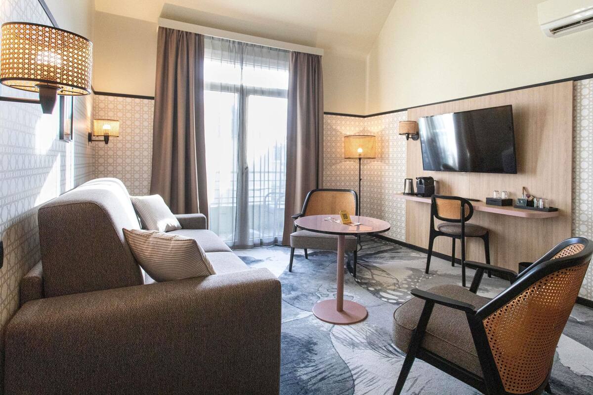 A hotel room with a sofa, a round table, a TV, and a window with curtains.