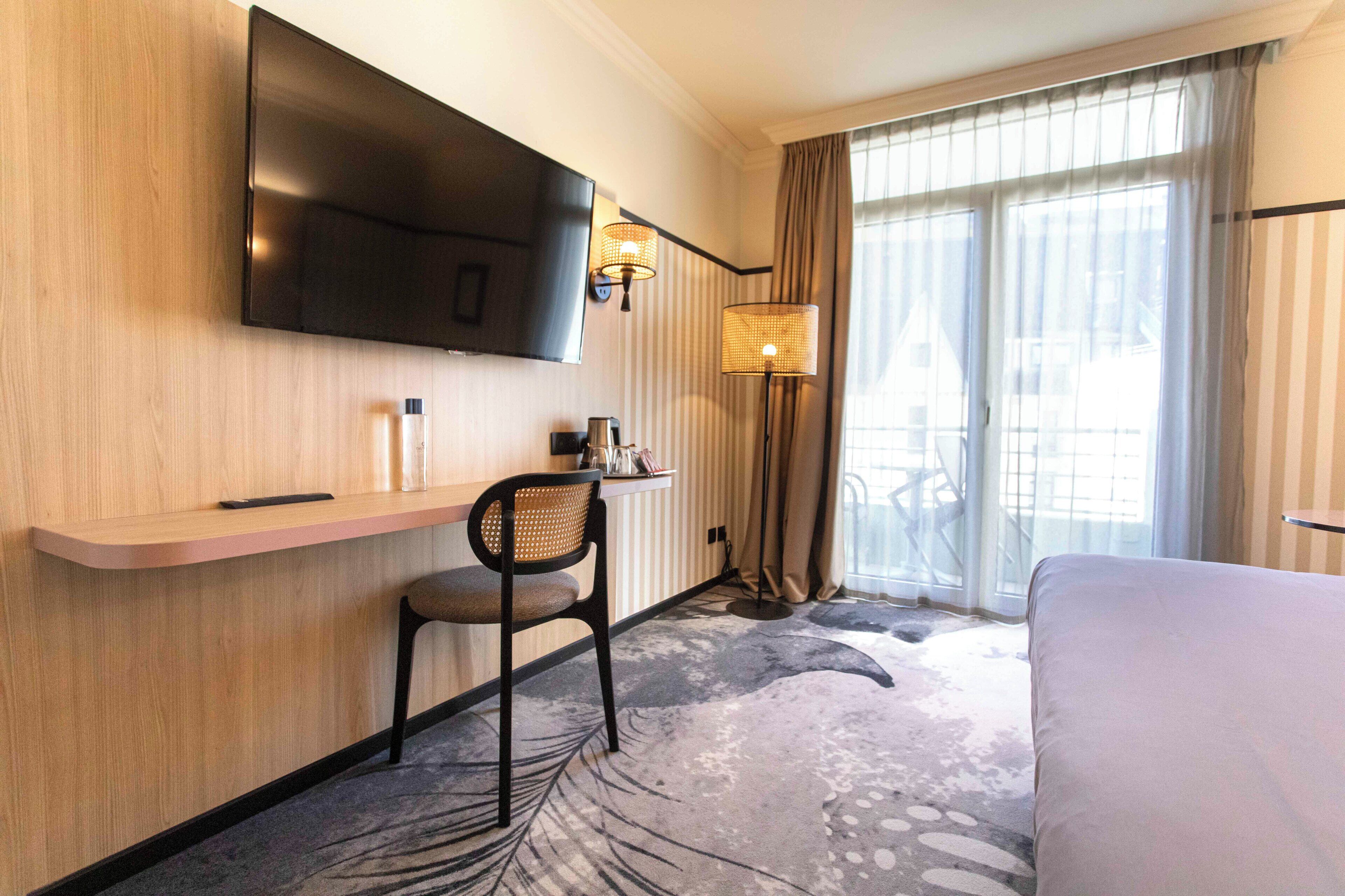 Habitación de hotel con televisor de pantalla plana en la pared, un escritorio con silla, una cama y un ventanal con cortinas.