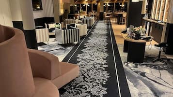 Um saguão de hotel com um tapete central em preto e branco, áreas de estar com cadeiras listradas e de cores sólidas, e uma recepção com balcão e objetos decorativos.