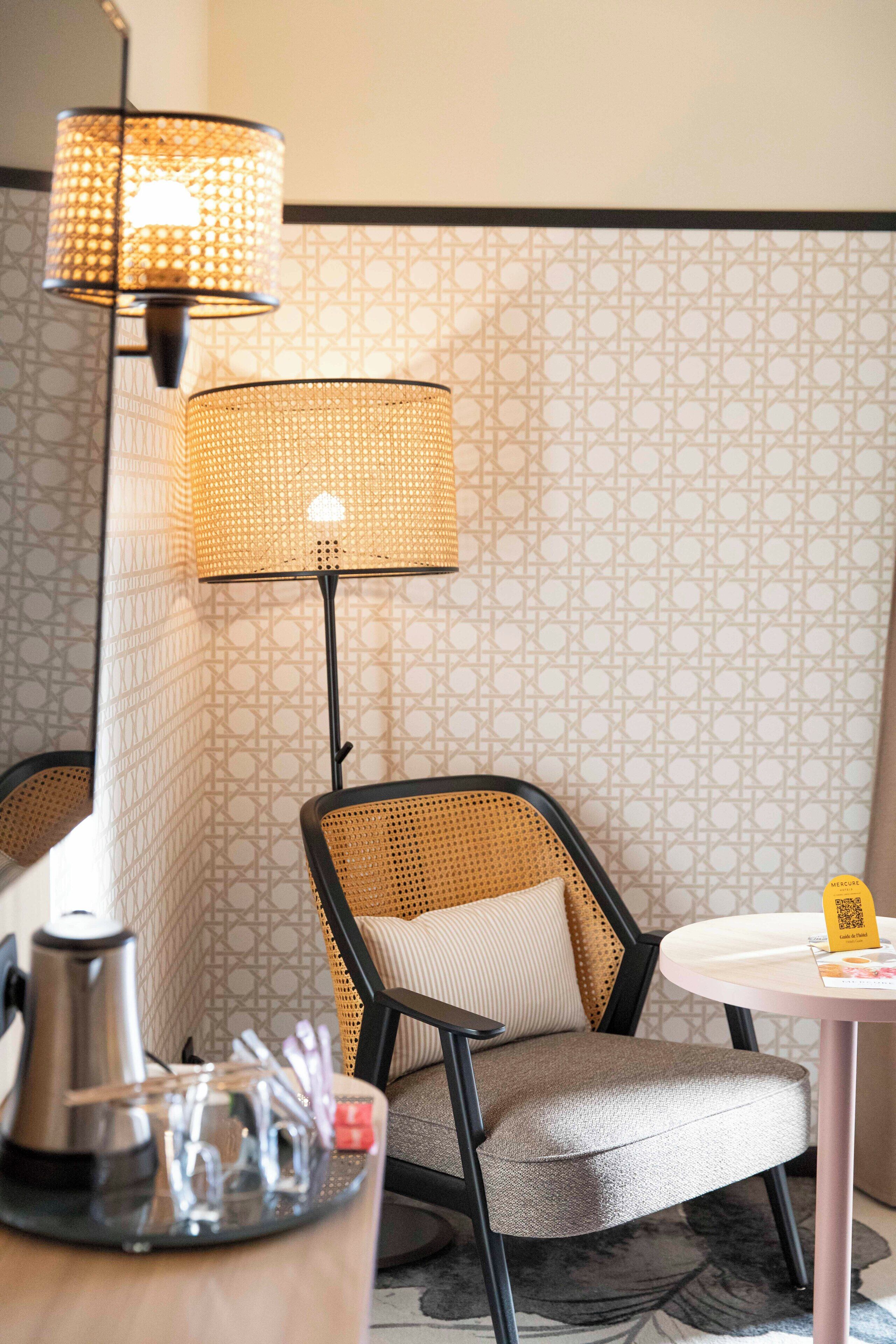 Une pièce avec un papier peint à motifs, une chaise en osier, une table dotée d’un code QR et un lampadaire.