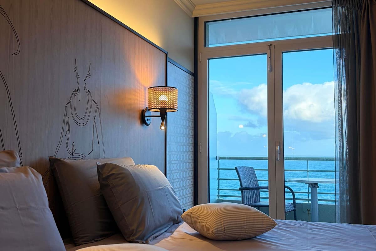 A hotel room with a large bed, a wall-mounted lamp, and a balcony with a view of the sea.