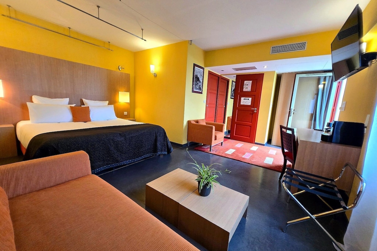 Ein Hotelzimmer mit Bett, einer Couch, einem Couchtisch mit Pflanze, einem Fernseher und einem Schreibtisch mit Stuhl.