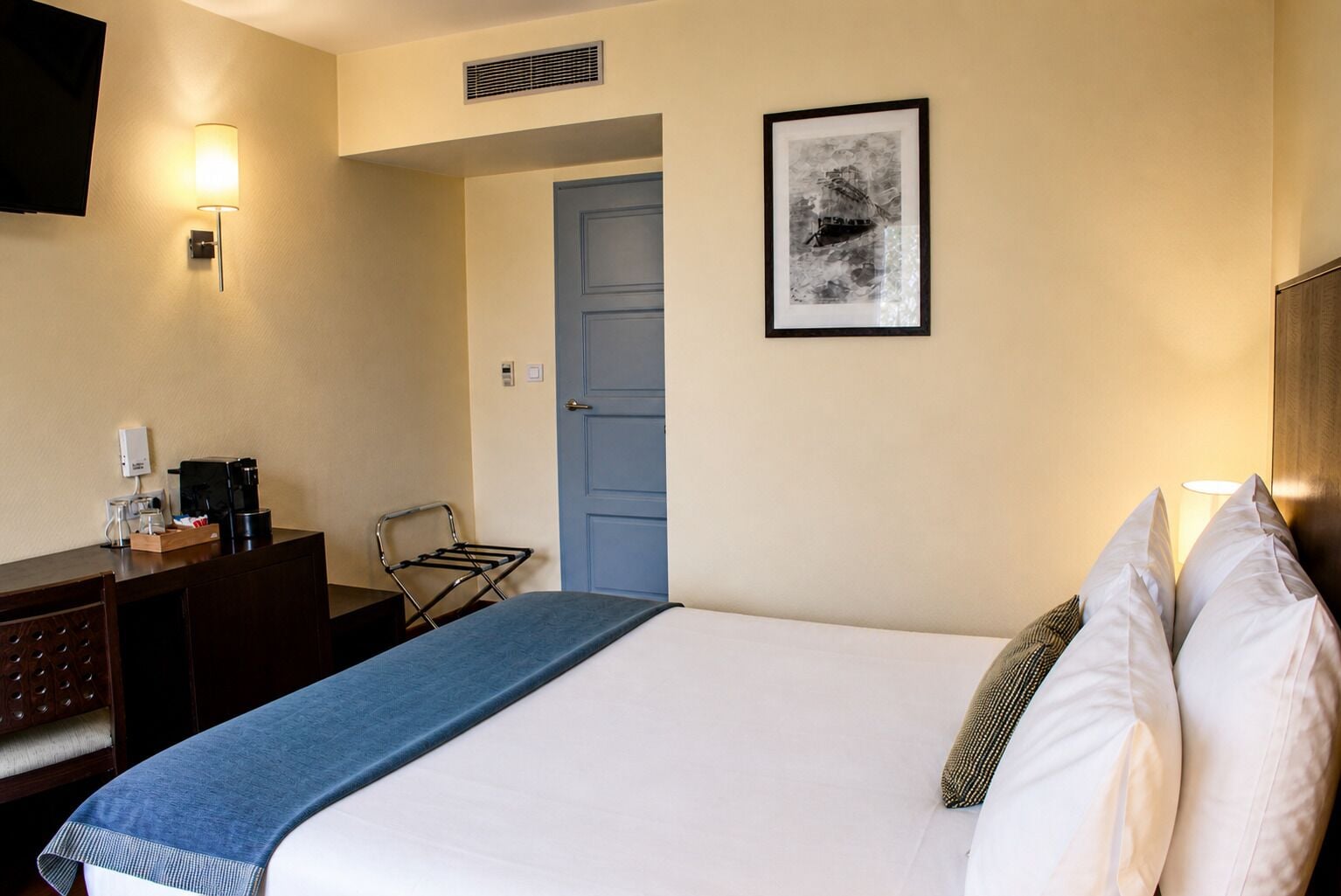 A hotel room with a bed, a desk with a coffee maker, a chair, a door, and a framed picture on the wall.