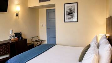 A hotel room with a bed, a desk with a coffee maker, a chair, a door, and a framed picture on the wall.