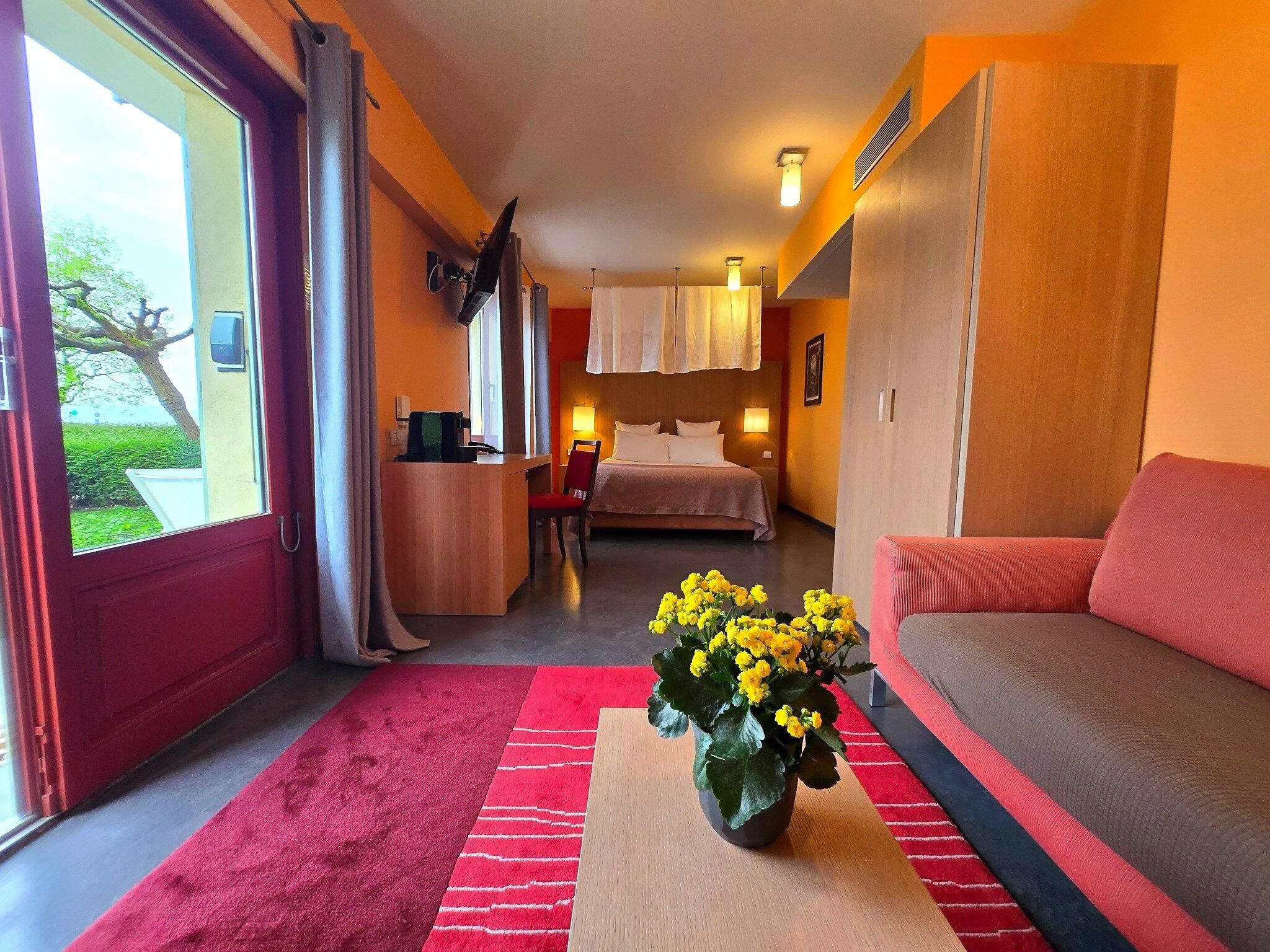 A hotel room with a bed, a desk, a chair, a sofa, a window with curtains, a vase with flowers, and a red carpet.