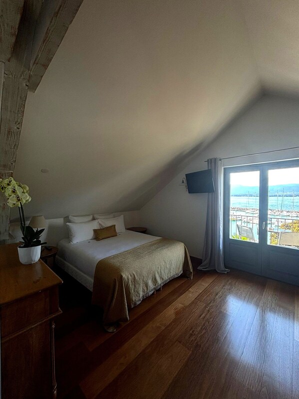 A bedroom with a bed, a potted plant, a desk, and a large window with a view of the sea. - La Riviera Victoria (Yvoire)