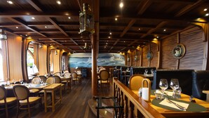 Restaurante com interiores em madeira, mesas com cadeiras e um mural de um navio na parede.