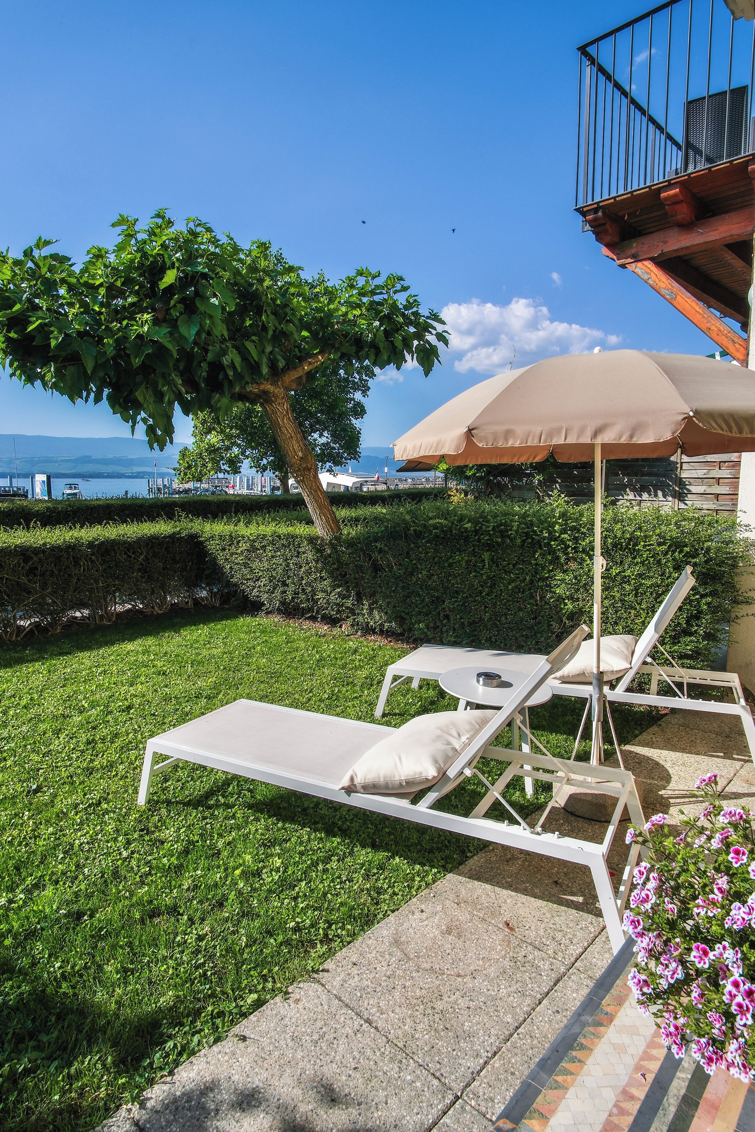 Une terrasse avec deux chaises longues blanches et un parasol beige, un arbre, et une vue sur la mer.