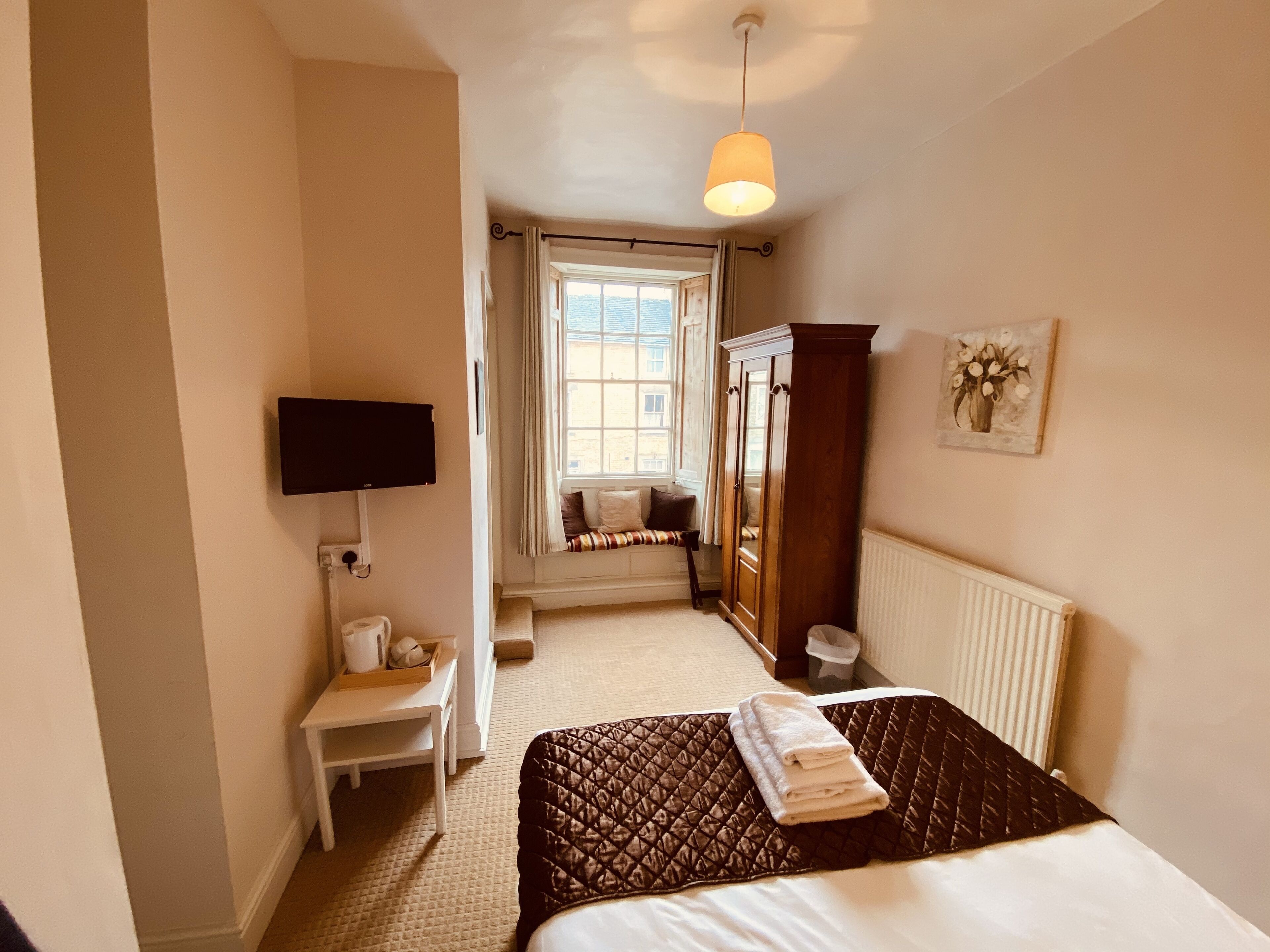 A bedroom with a bed, a television mounted on the wall, a window with curtains, a radiator, and a wooden wardrobe.