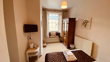 Een slaapkamer met een bed, een aan de muur bevestigde televisie, een raam met gordijnen, een radiator en een houten kledingkast.