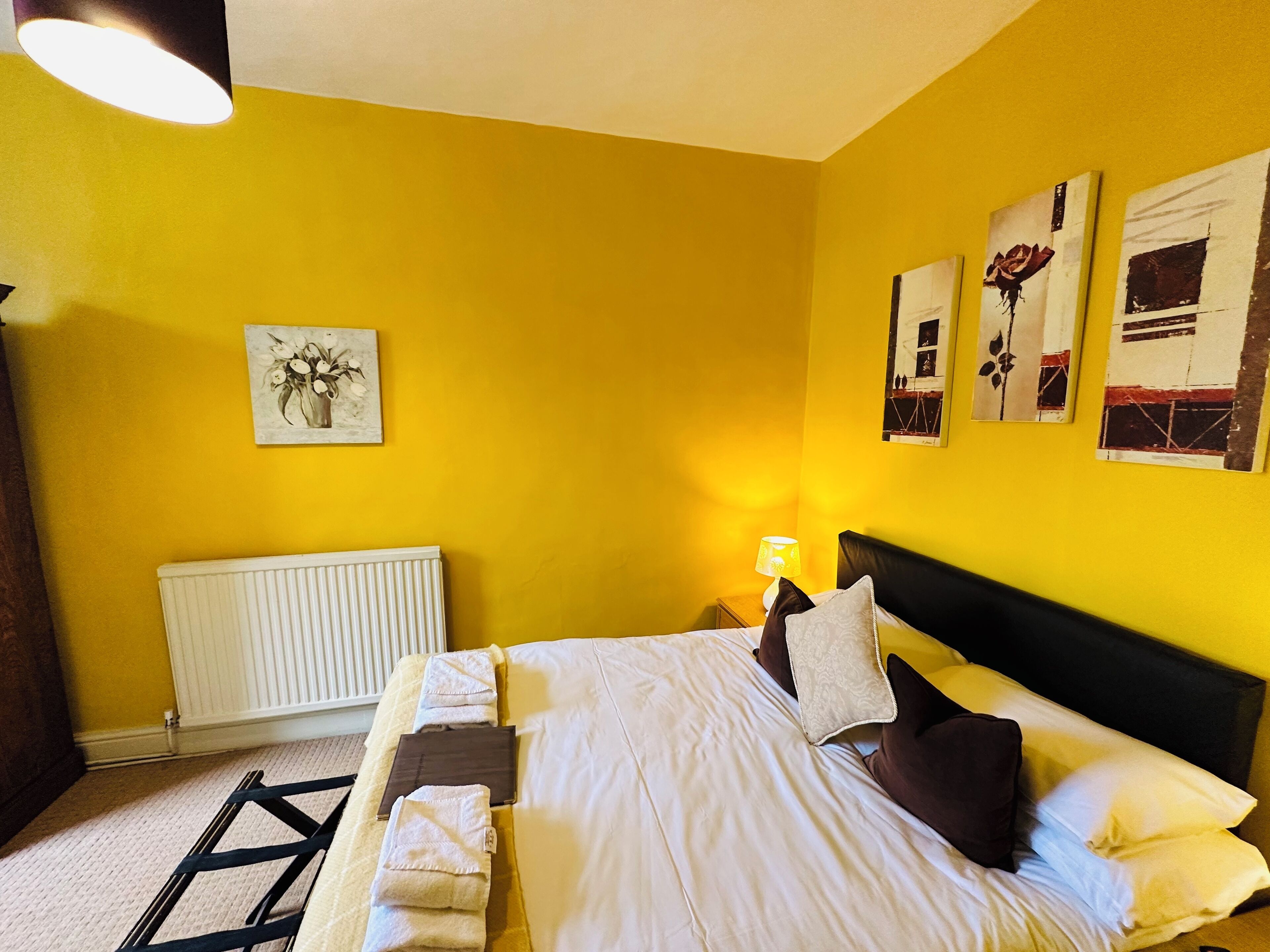 A hotel room with a yellow wall, a bed with white bedding, a radiator, and artwork on the walls.