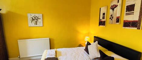 A hotel room with a yellow wall, a bed with white bedding, a radiator, and artwork on the walls.