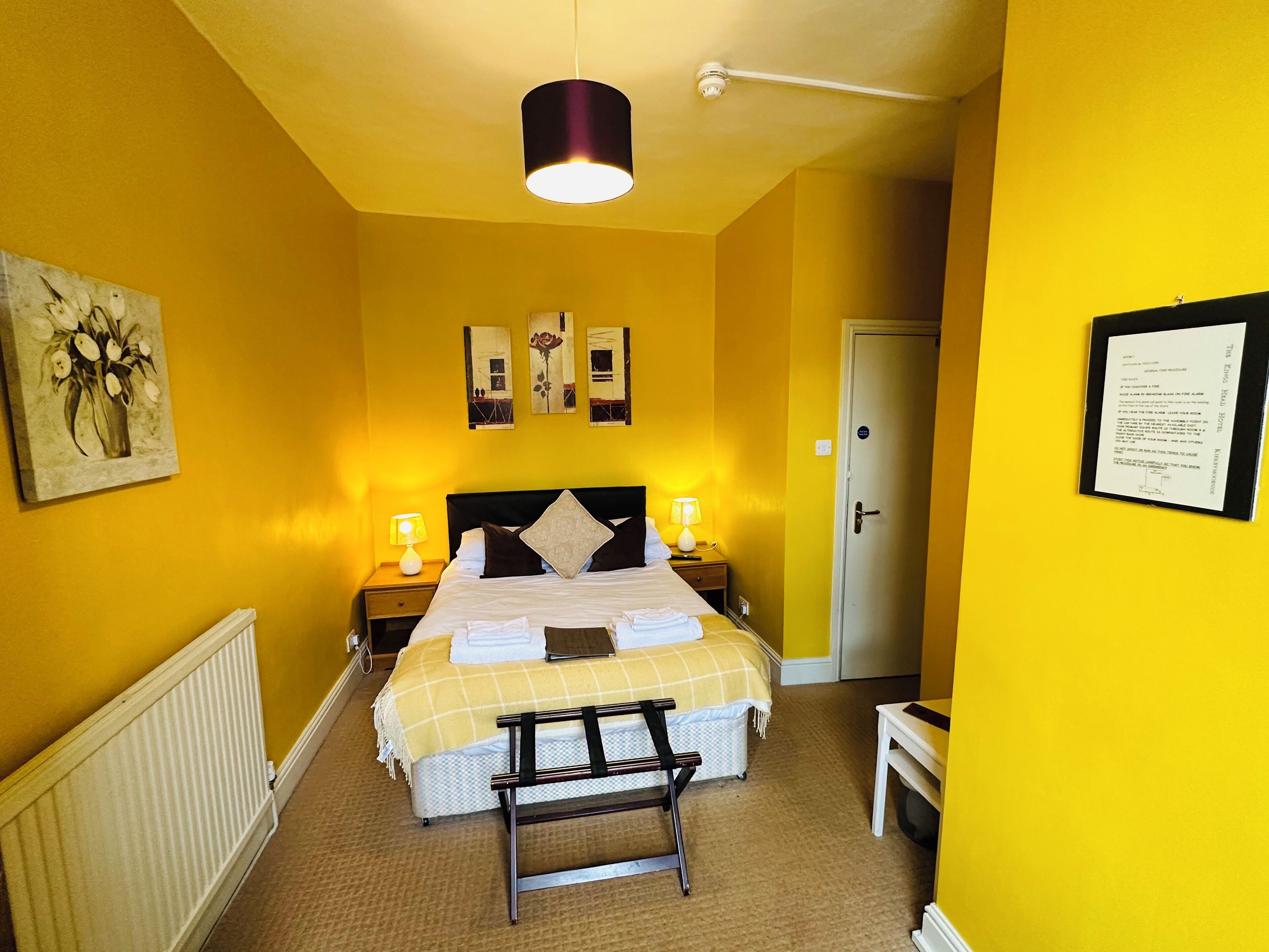 Een hotelkamer met een bed, nachtlampen, een bankje, een radiator en een deur.