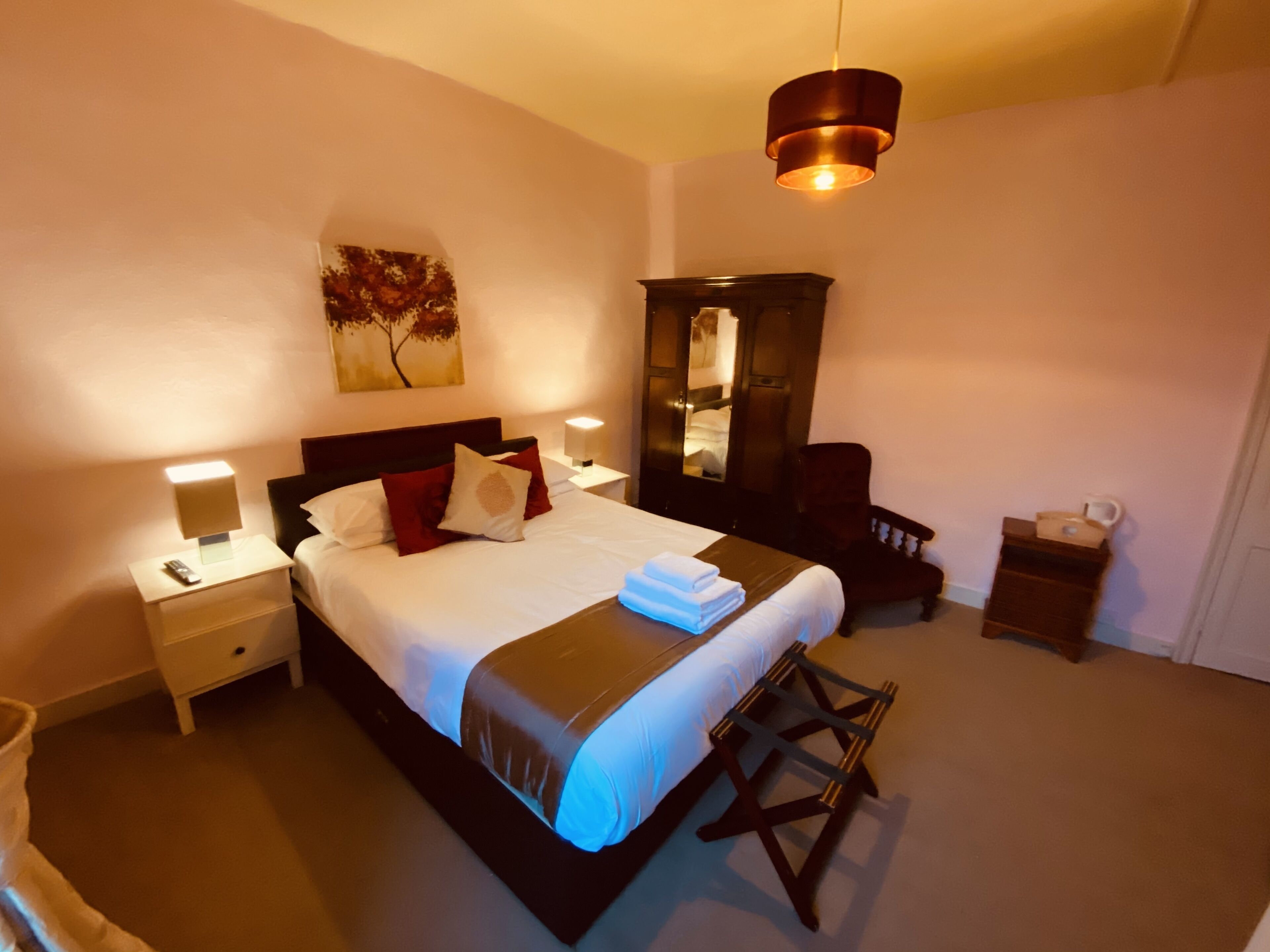 A neatly made bed with white linens and red pillows, a bedside table with a lamp, a chair, a small table with a cup, and a painting on the wall.