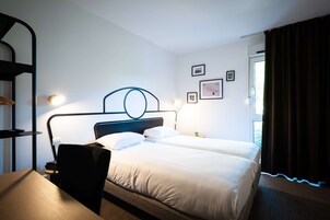 Une chambre d’hôtel avec un lit double, un bureau, une chaise, une table de chevet, une lampe et une fenêtre avec des rideaux.