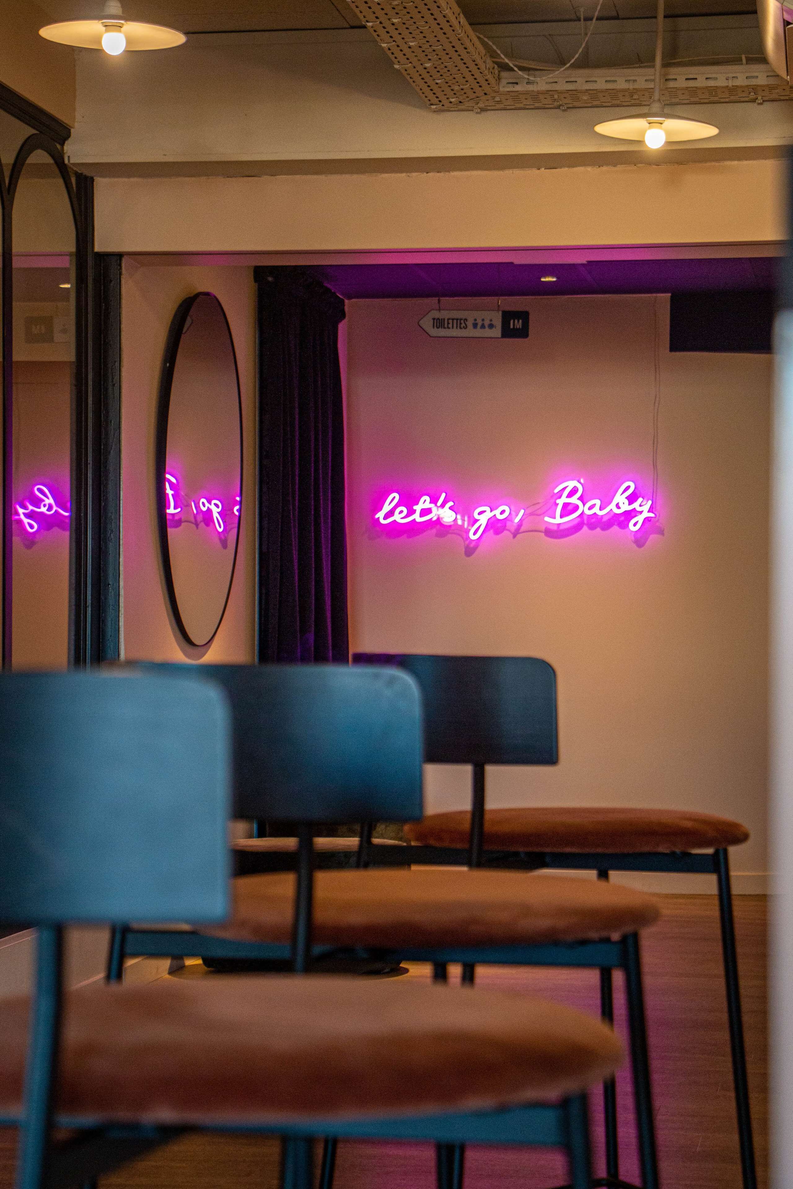 Una stanza con un neon che riporta la scritta 'Let's go, Baby' e un'indicazione per il bagno maschile. Sono presenti due sedie con cuscini marroni e struttura nera.