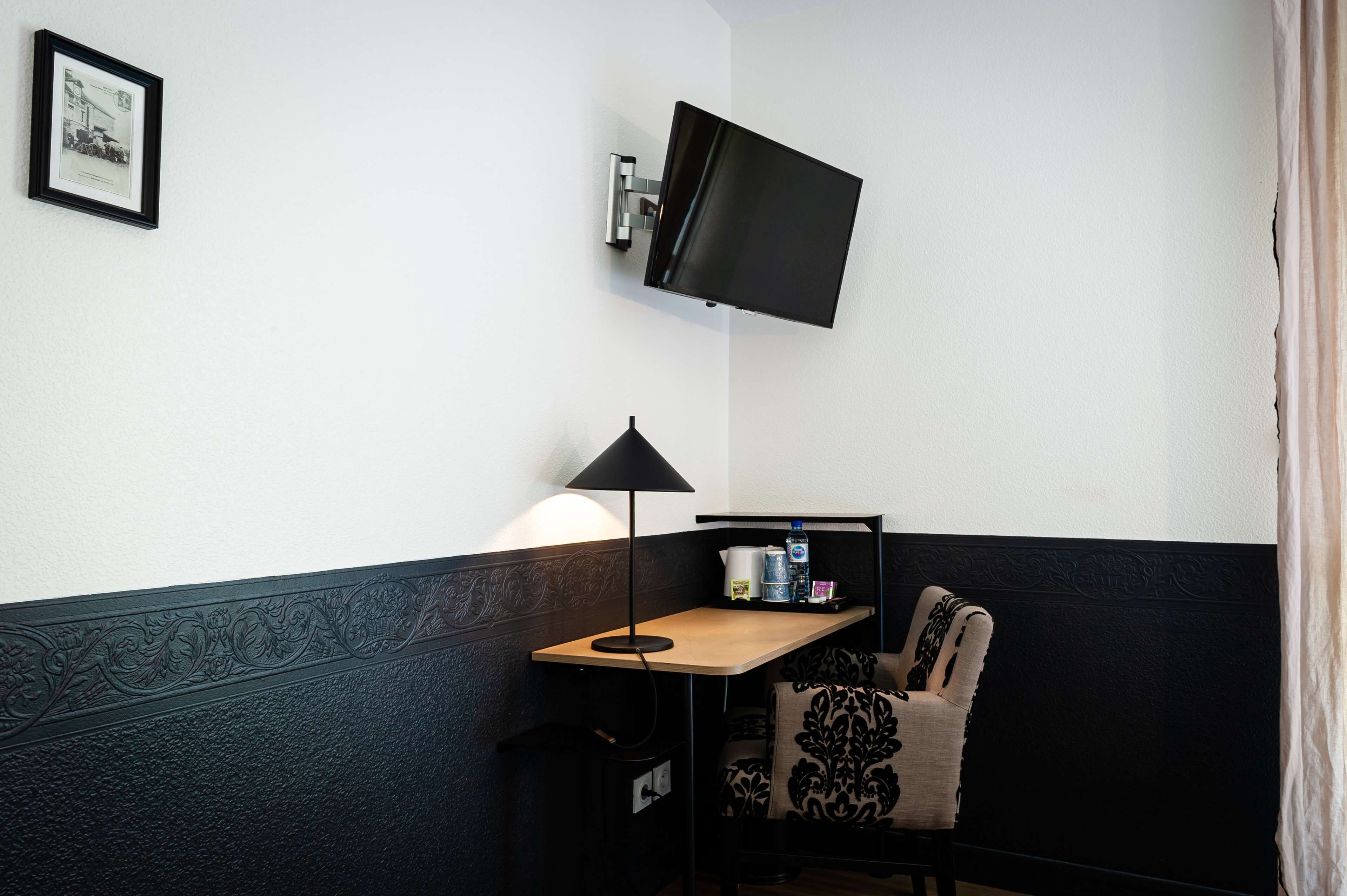 A corner room with a flat-screen TV mounted on the wall, a small table with a lamp, and a chair with a patterned cushion.