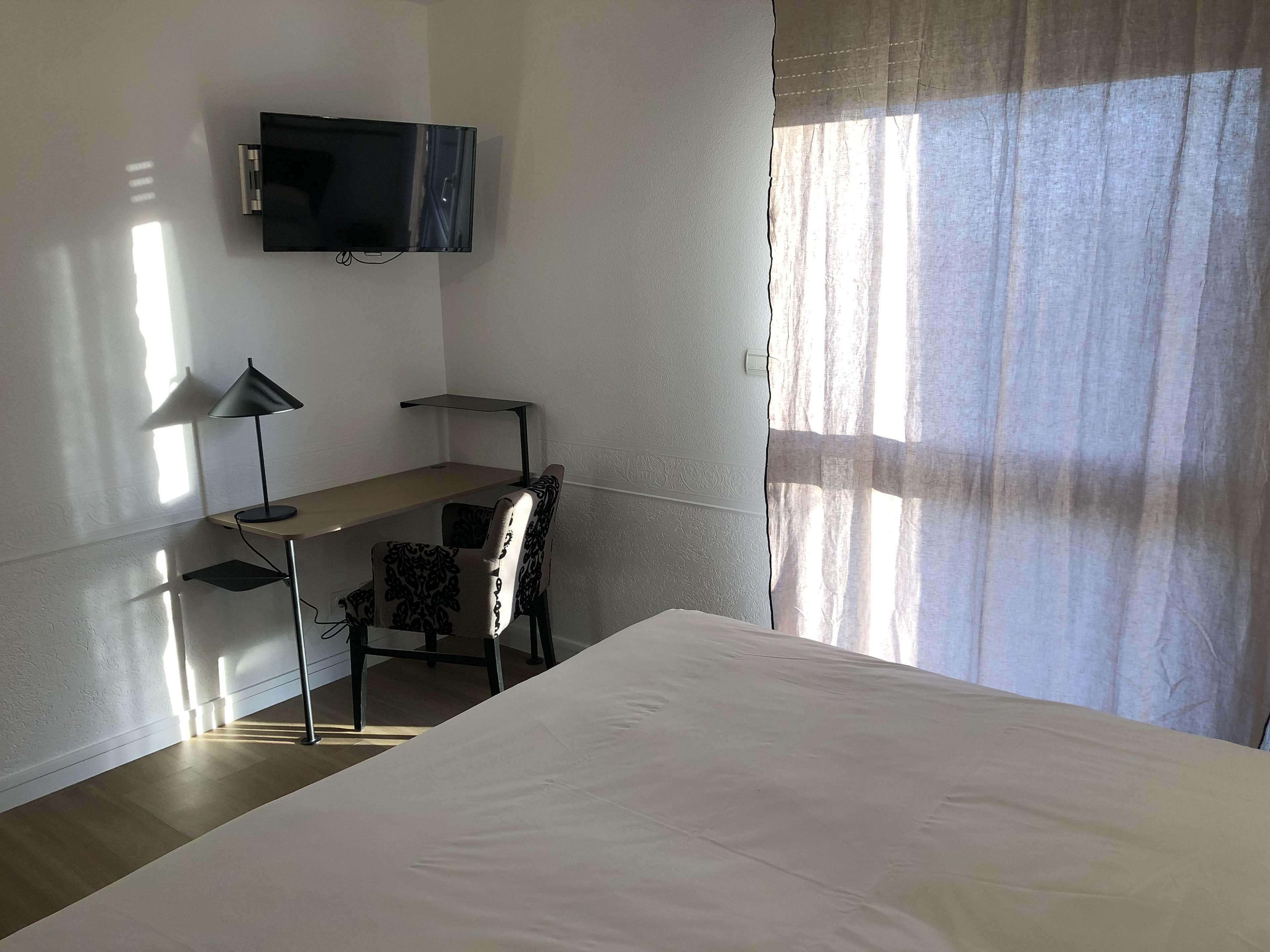 A hotel room with a bed, a desk with a chair, a TV mounted on the wall, and a window with curtains.