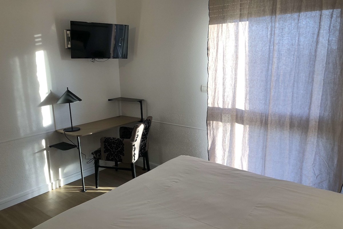 Habitación de hotel con cama, escritorio con silla, televisor en la pared y ventana con cortinas.