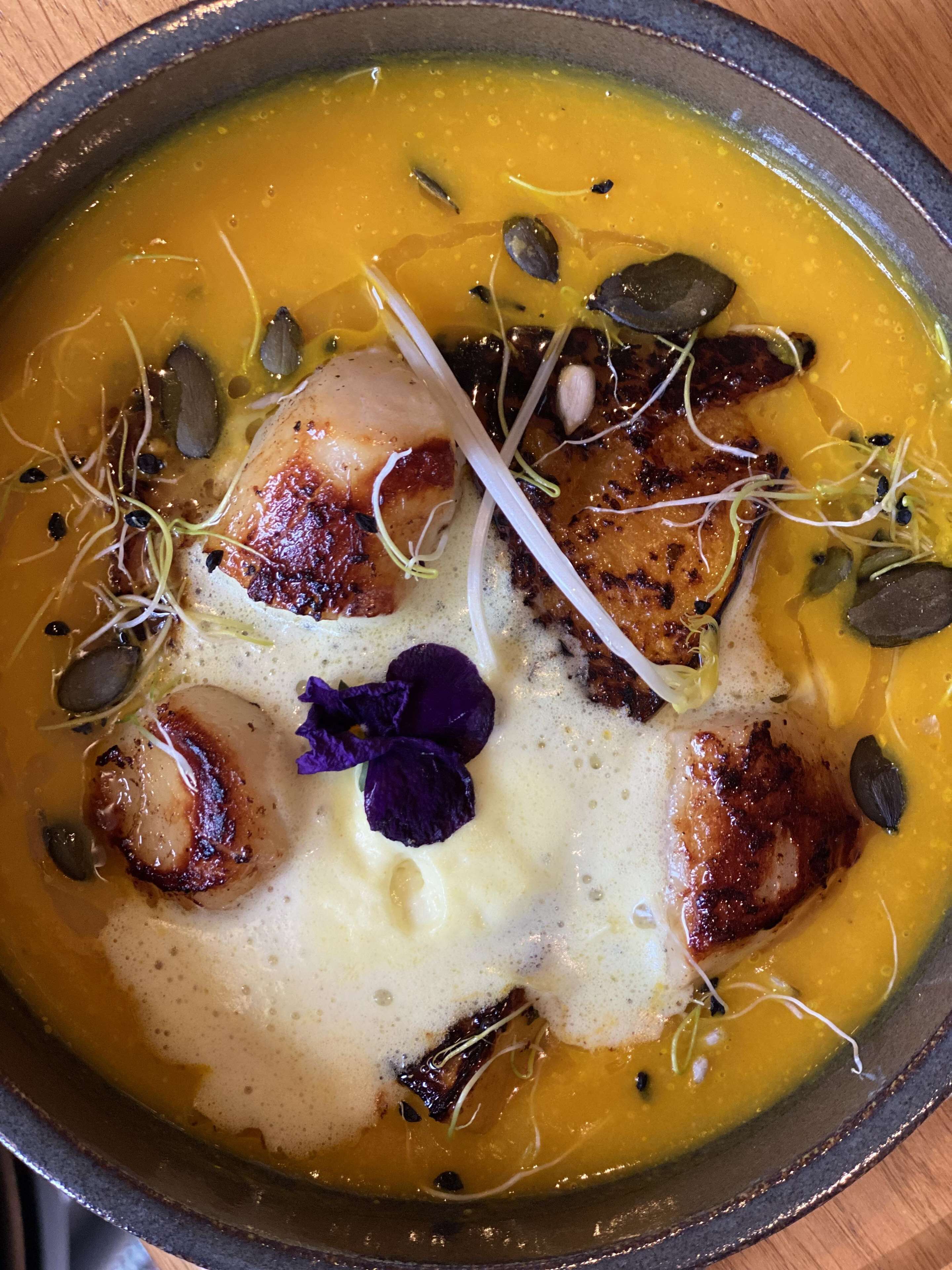 Un tazón de sopa con vieiras selladas, semillas de calabaza y brotes, adornado con flores moradas.
