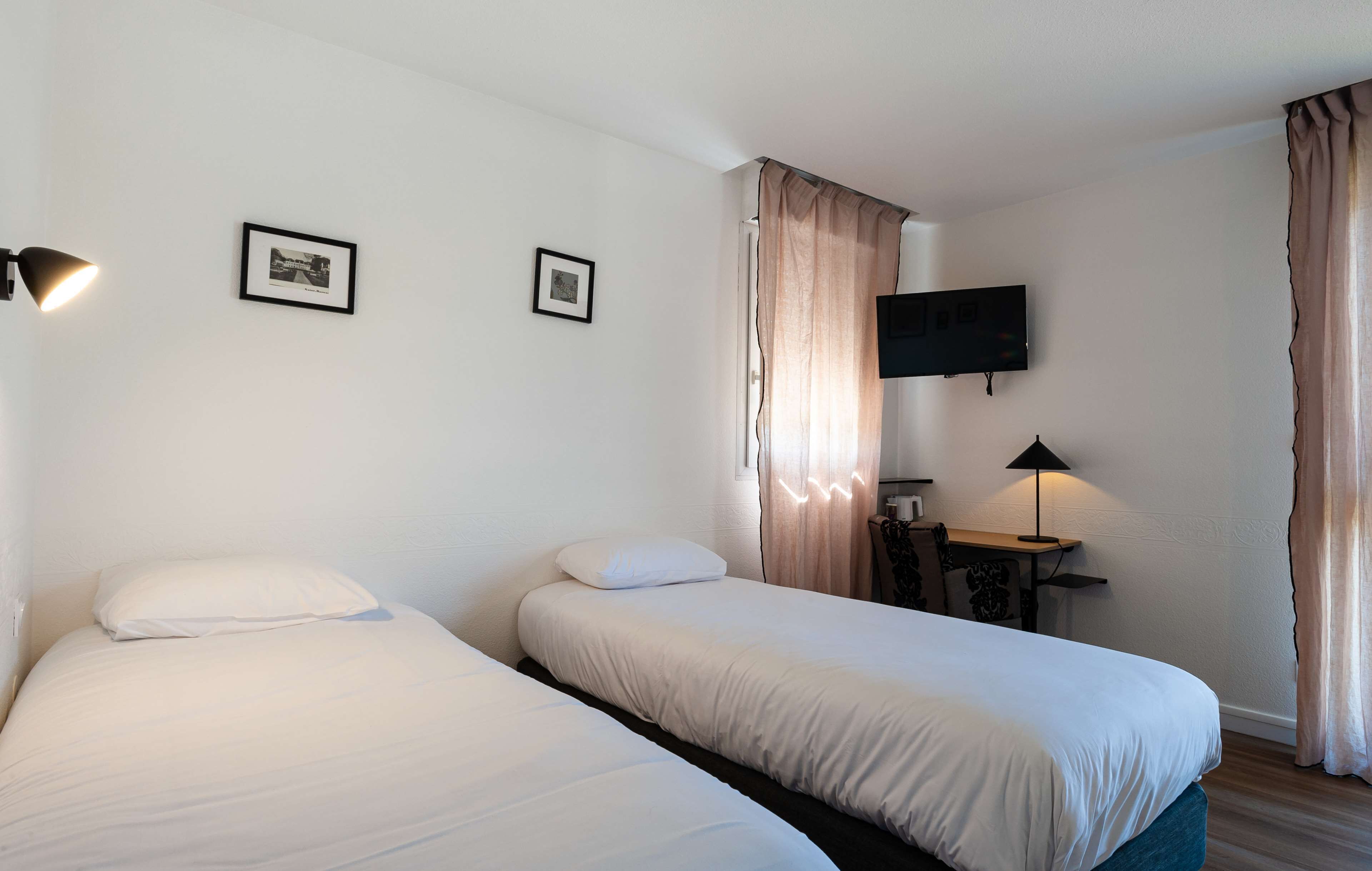 A hotel room with two beds, a desk, a chair, a TV mounted on the wall, and a lamp.