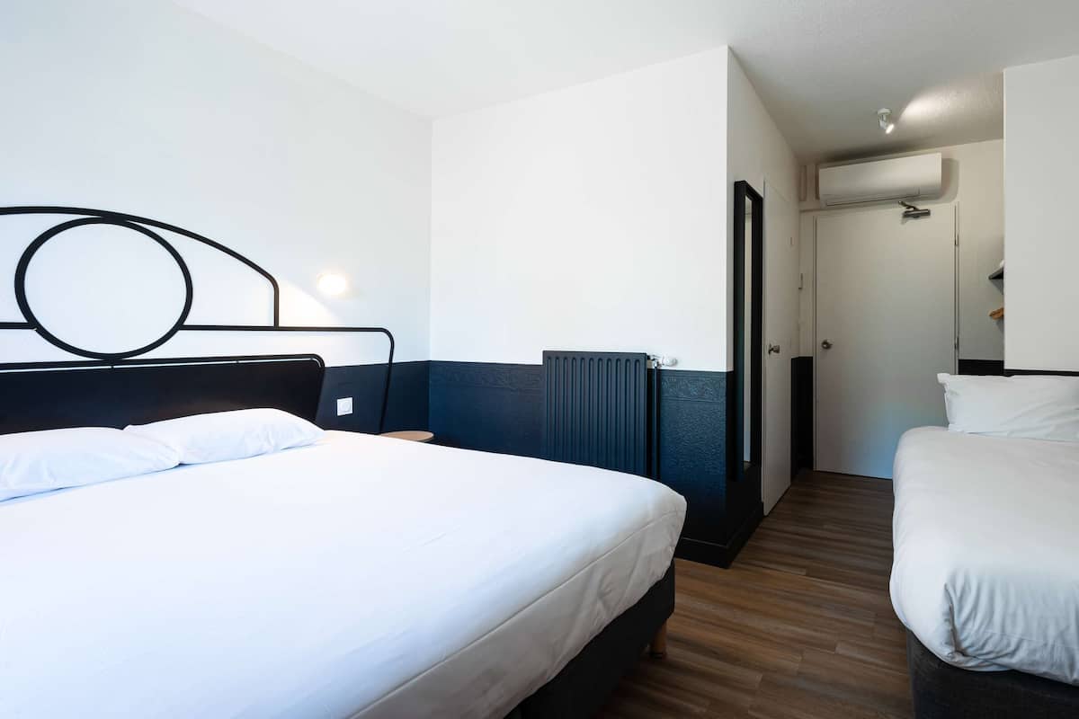 Una habitación de hotel con una cama grande, una cama más pequeña, una mesita de noche, un aire acondicionado instalado en la pared y una puerta que da a otra habitación.