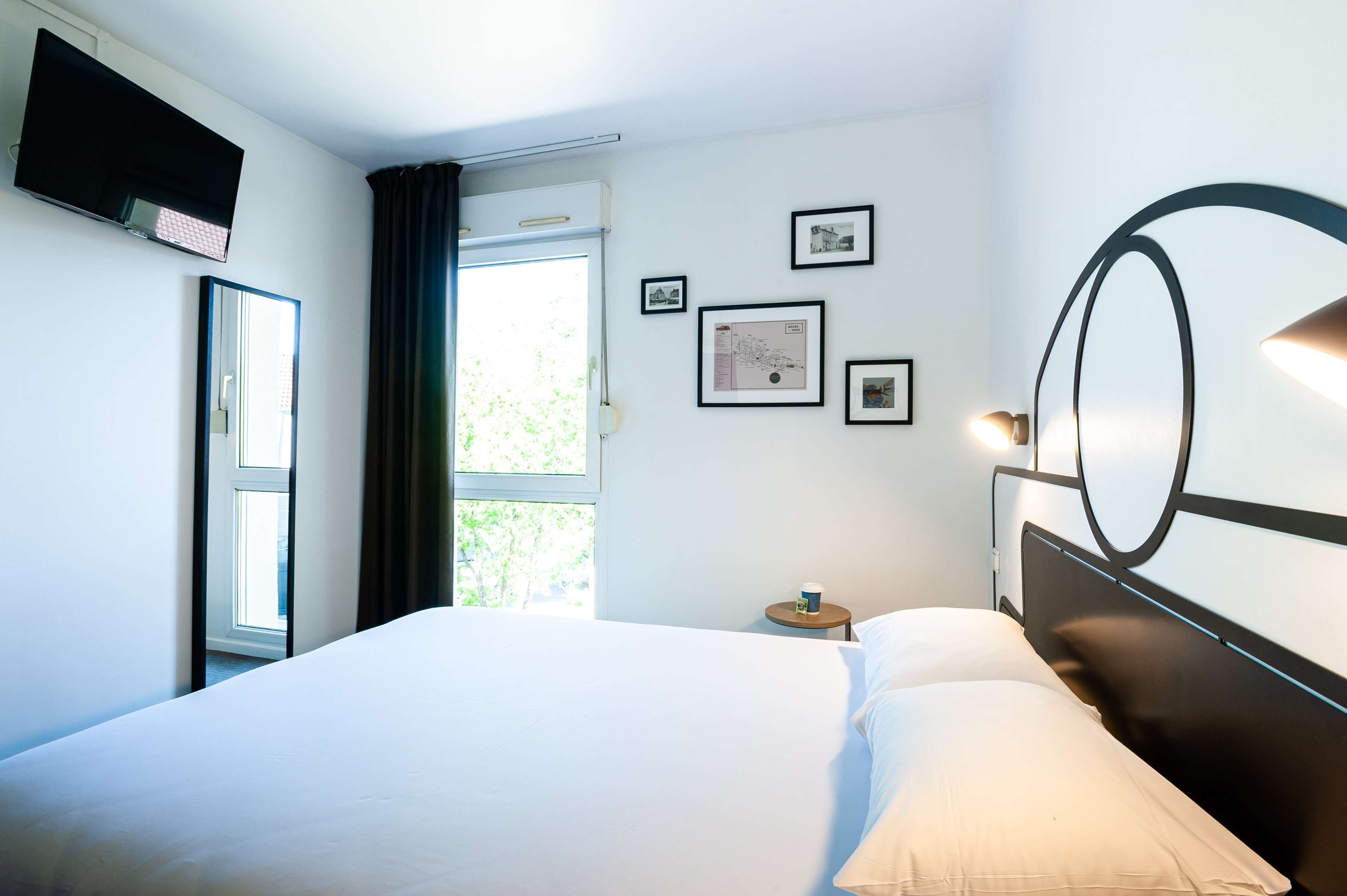 A hotel room with a bed, a flat-screen TV mounted on the wall, a small table with a cup, and framed pictures on the wall.