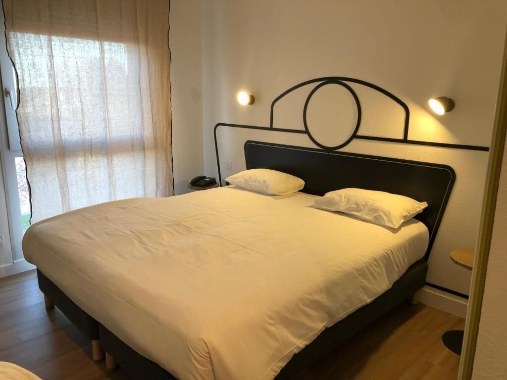 A neatly made bed with a black metal headboard and two pillows, a bedside table with a lamp, and a window with sheer curtains.