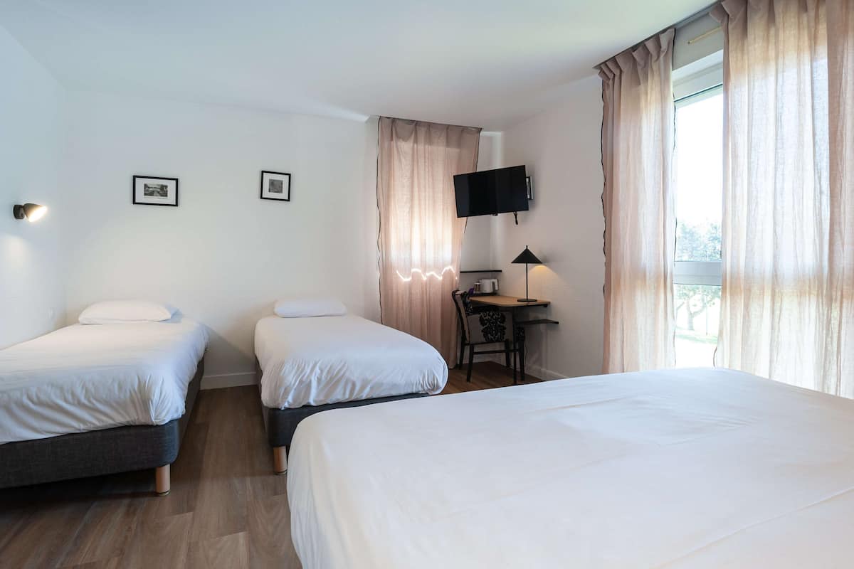 Habitación de hotel con dos camas individuales, un escritorio con silla, un televisor de pantalla plana en la pared y una ventana con cortinas.