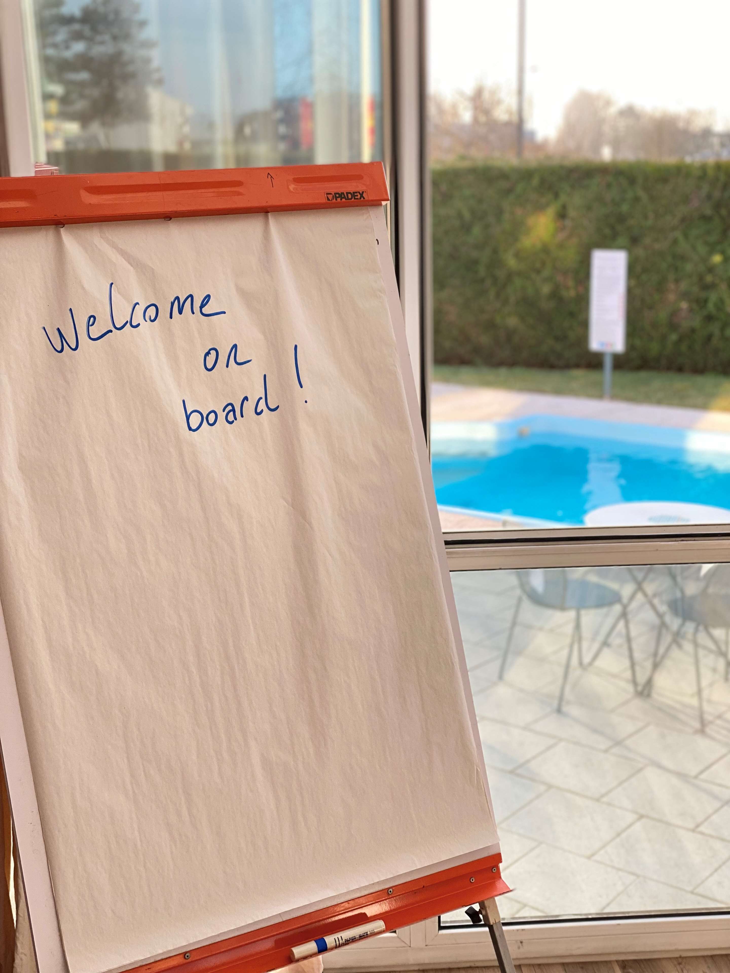 A whiteboard with "Welcome on board!" written on it, placed near a window overlooking a swimming pool.