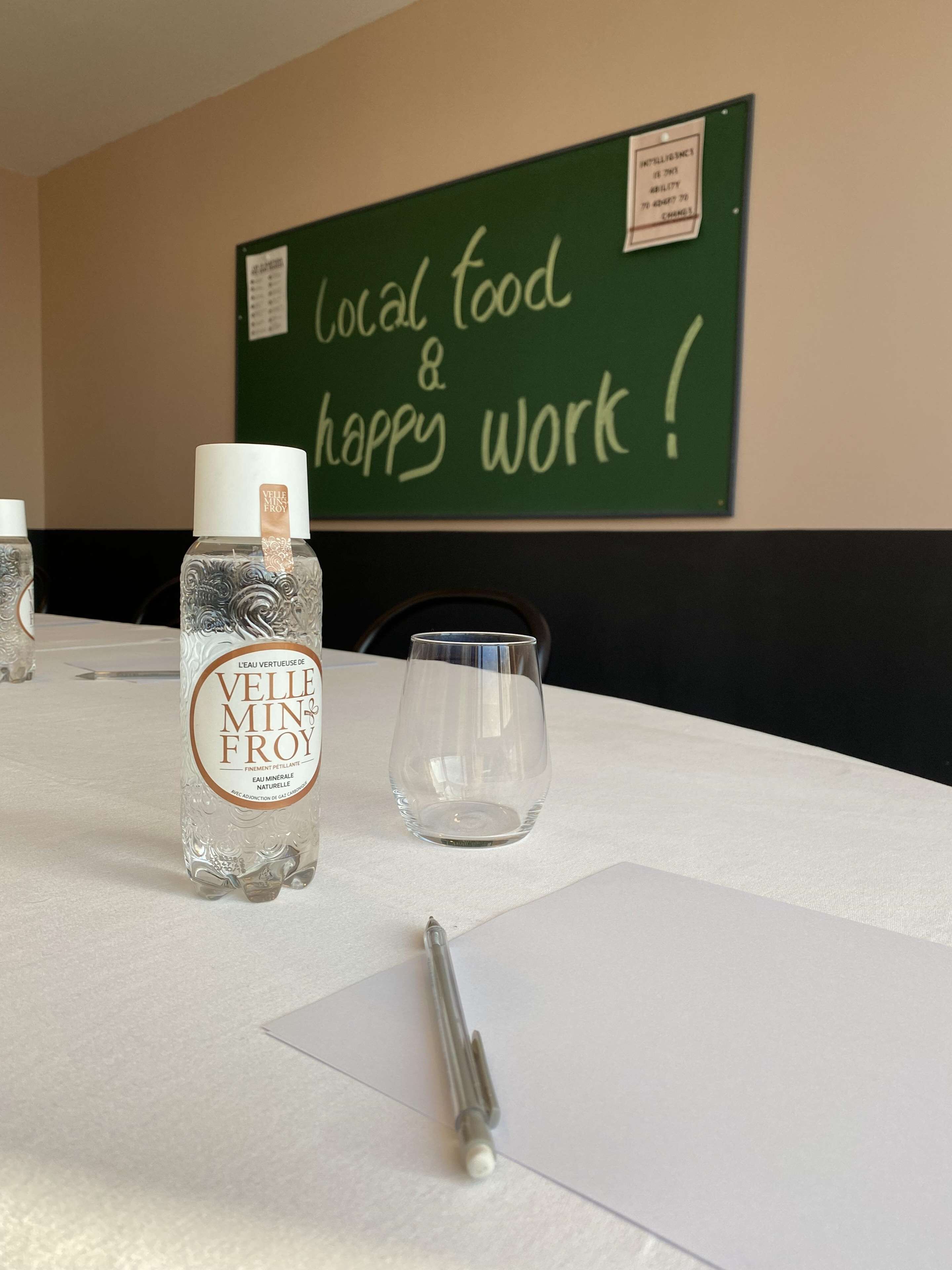 Une table avec une bouteille d’eau, un verre et un stylo. Un tableau noir en arrière-plan porte l’inscription : « Produits locaux et ambiance conviviale ! »