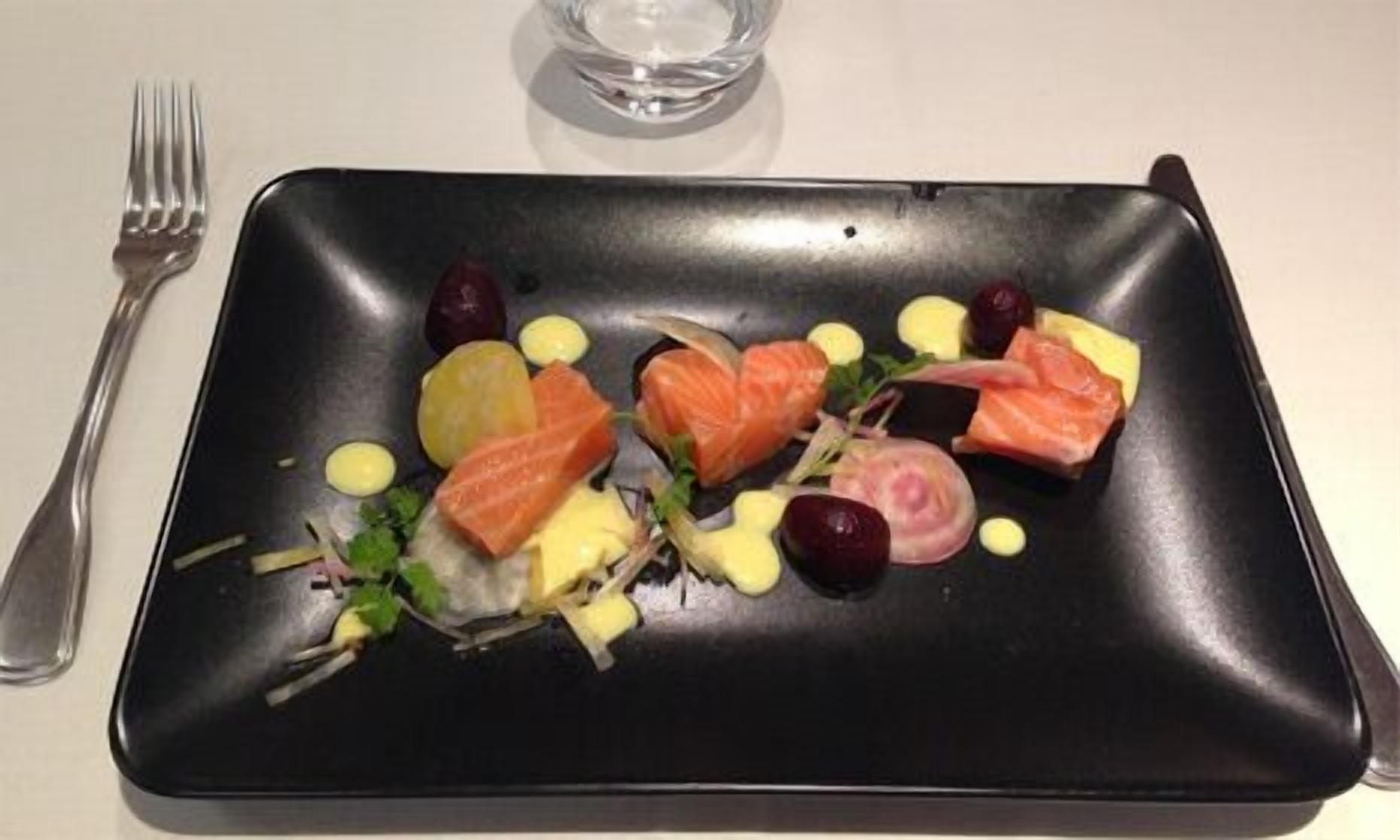 Plat negre rectangular amb diferents aliments, com salmó, raïm i verdures, acompanyat d'una forquilla a l'esquerra.