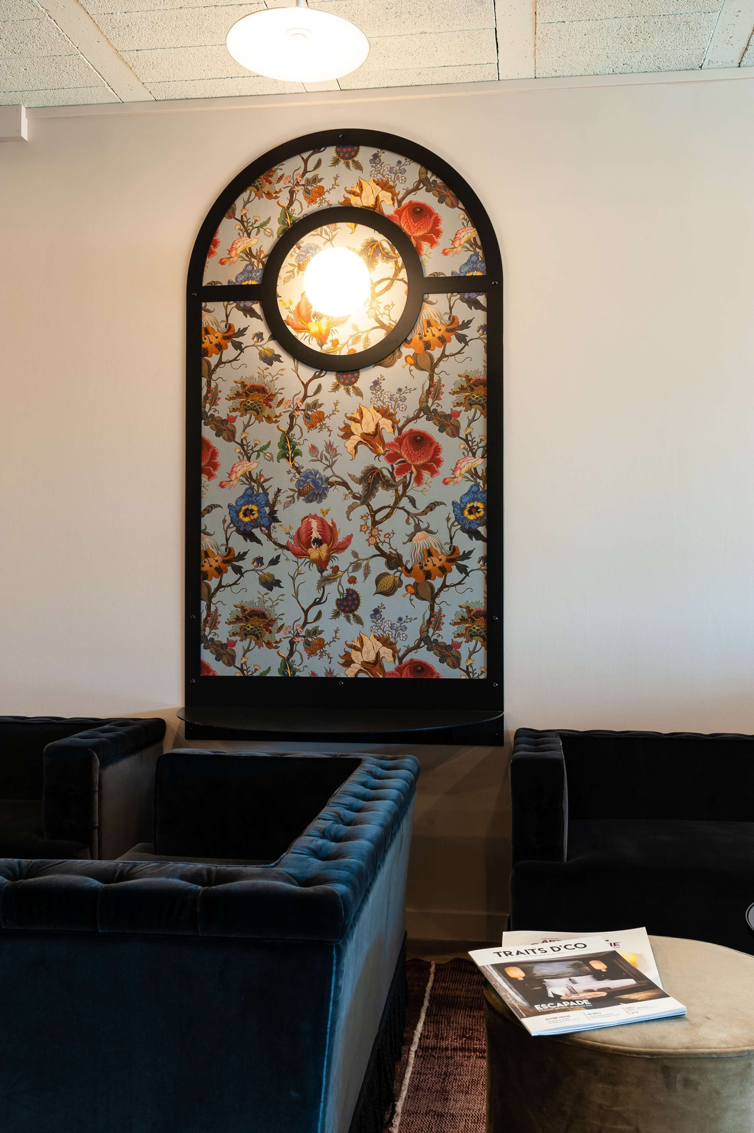 Una habitación con un ventanal de gran tamaño con estampado floral, una zona de estar con cojines y una revista sobre una mesita.