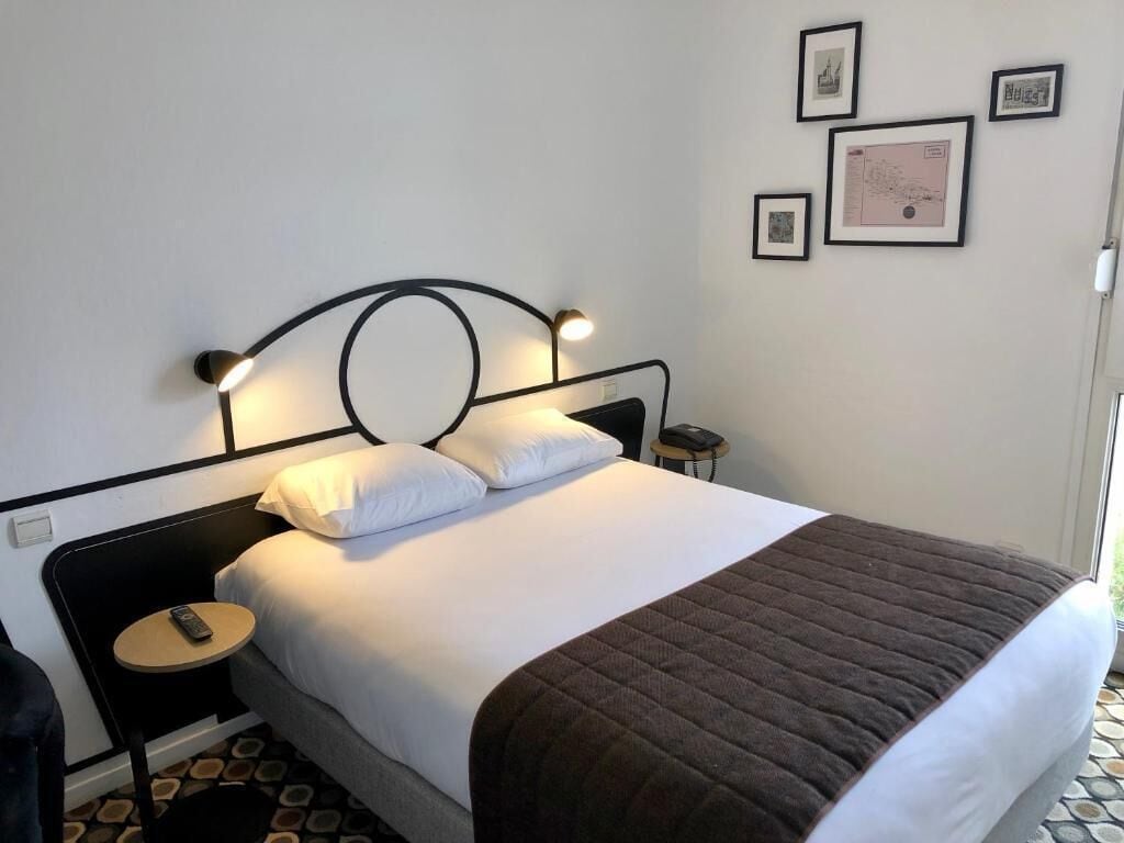 Una cama bien hecha con una manta marrón, una mesita de noche con una lámpara y una pared adornada con cuadros.