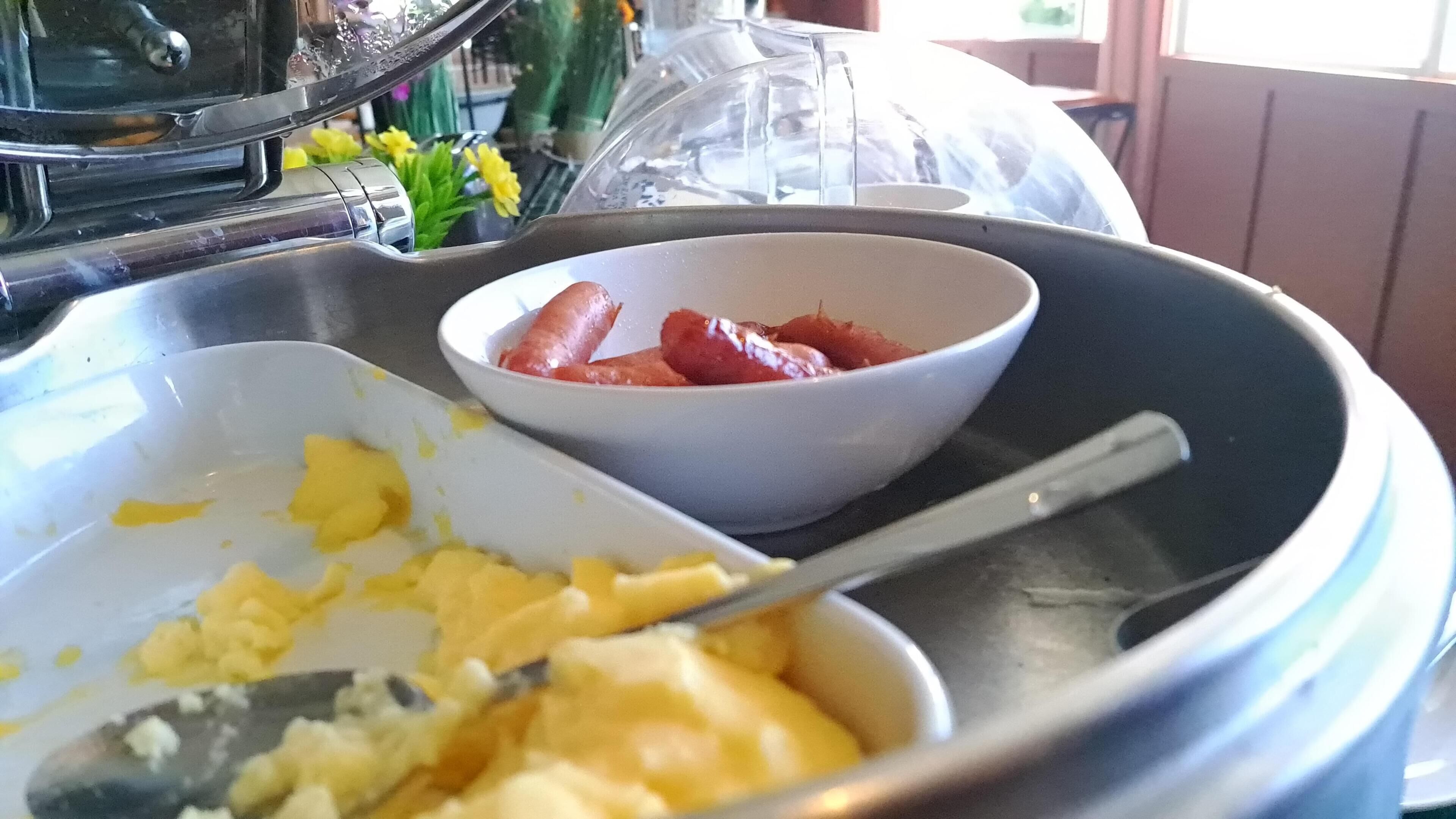 Un bufé de desayuno con huevos revueltos, salchichas y un bol de fruta.