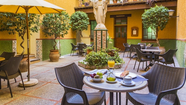 An outdoor patio with wicker chairs, a table with a drink and fruit, and a statue in the background. - Hostal Loreto (Denia)