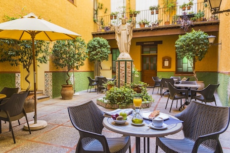 An outdoor patio with wicker chairs, a table with a drink and fruit, and a statue in the background.