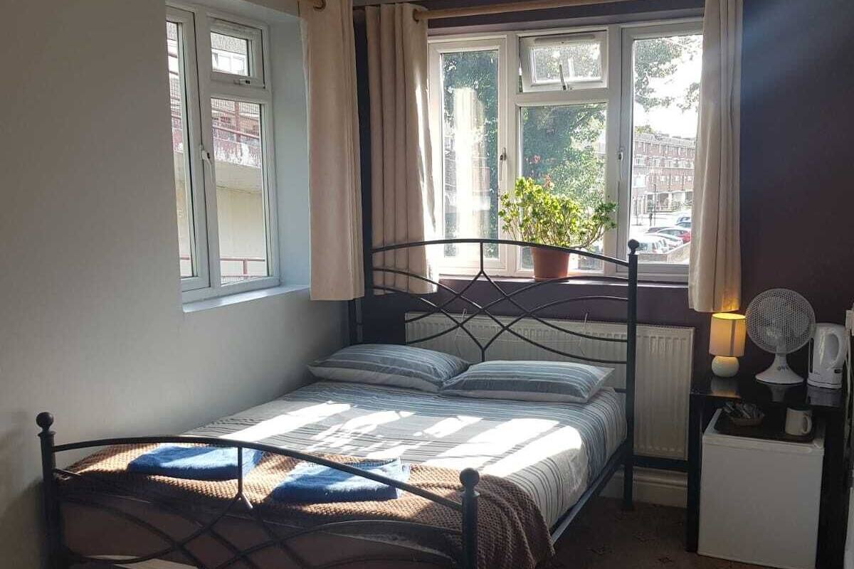 A small, well-lit bedroom with a single bed, a window with curtains, a potted plant, a bedside table with a lamp, and a radiator.