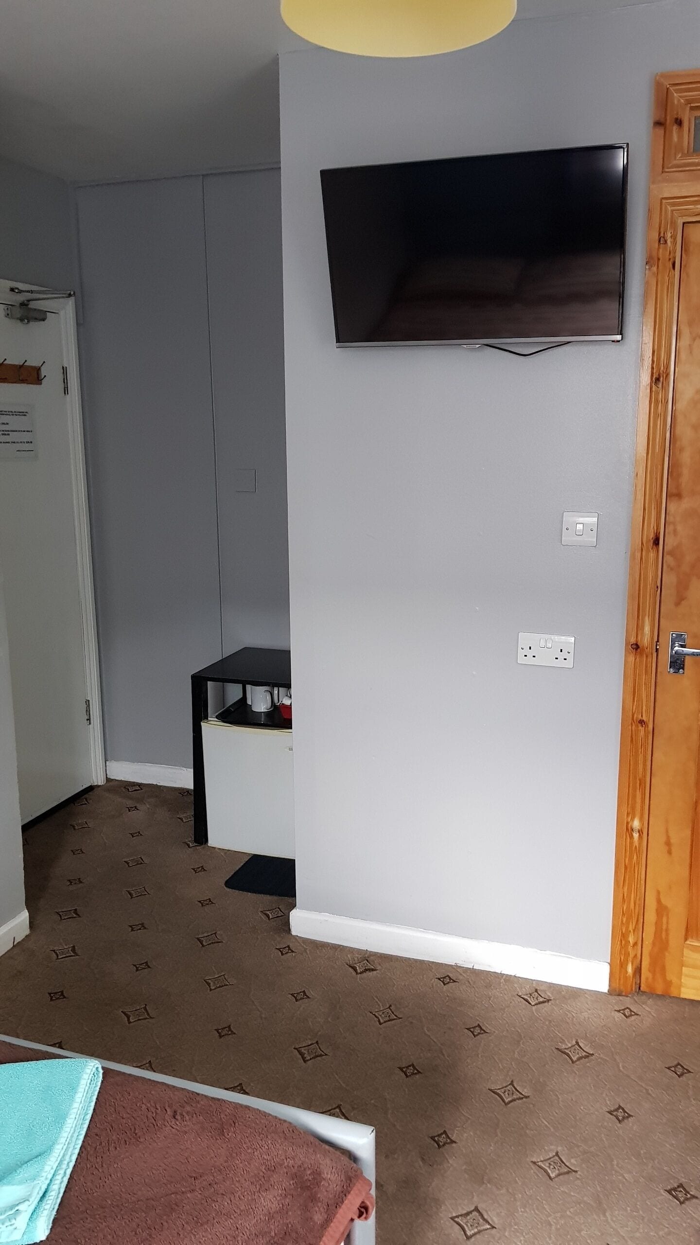 A room with a flat-screen TV mounted on the wall, a wooden door, a white door, a small black table with a kettle, and a patterned carpet.