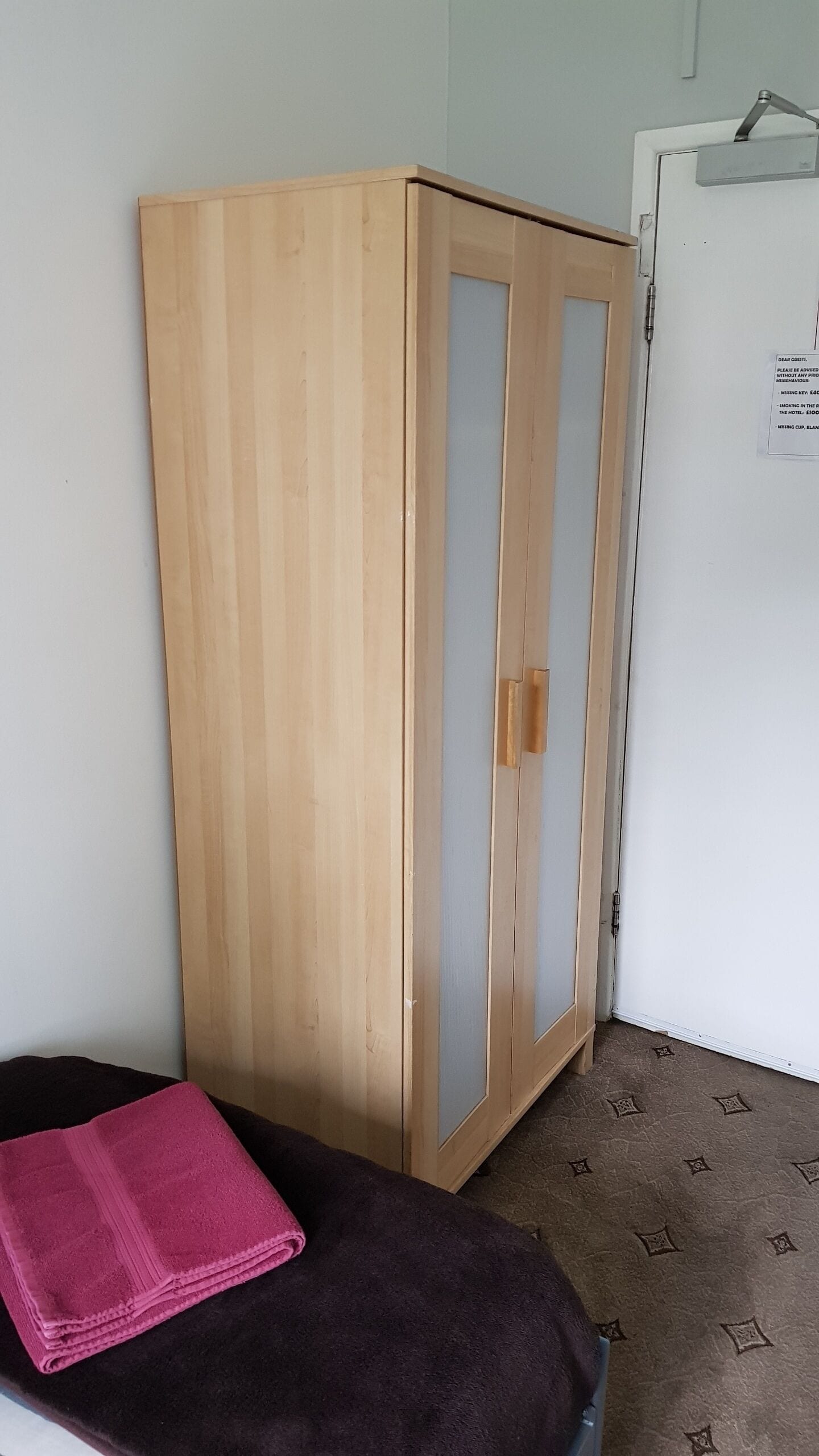 Una habitación con un armario de madera, una cama con una manta rosa y una puerta blanca con un cartel.