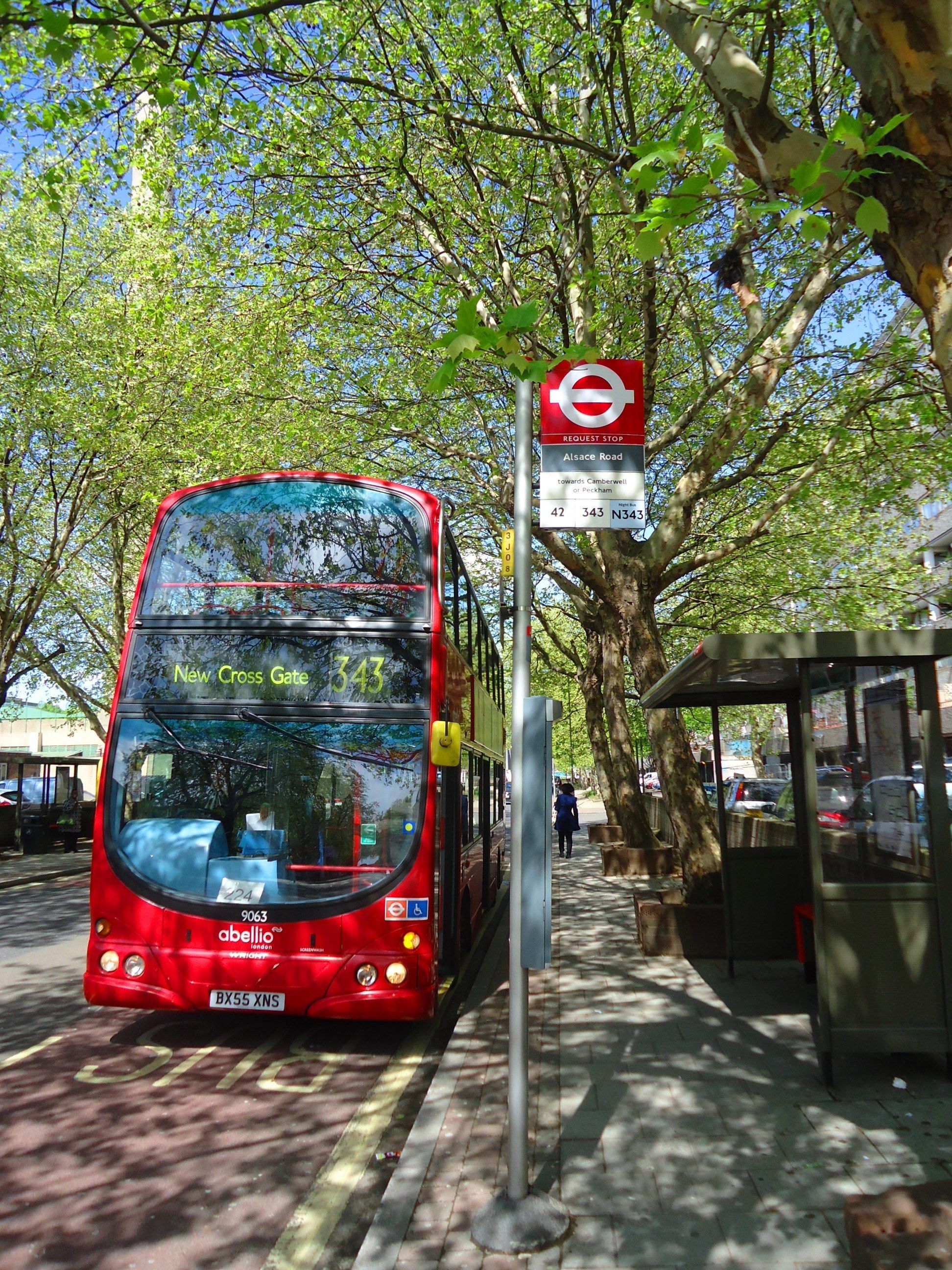 A red double-decker bus at a bus stop with a sign indicating Alsace Road and New Cross Gate.