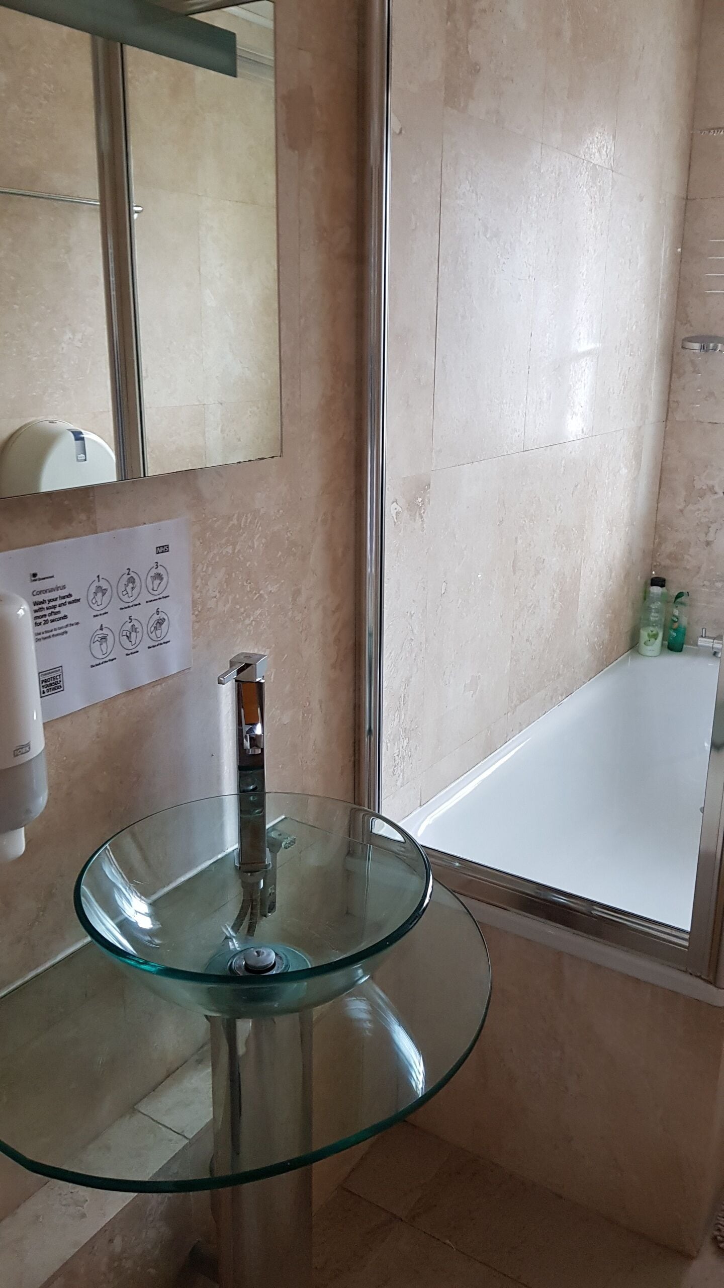 A bathroom with a glass sink, a mirror, a hand sanitizer dispenser, and a white bathtub.